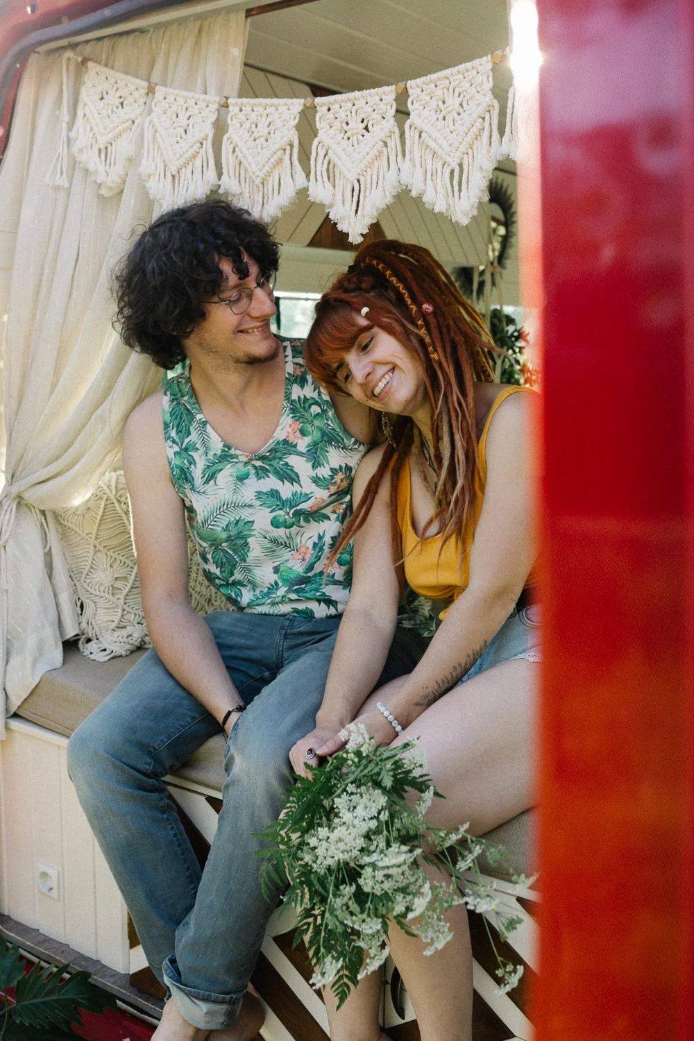 Zwei Menschen sitzen in einem Boho-inspirierten Raum mit Makramée-Gardinen und dekorativen Stoffen, lachen und verbringen eine fröhliche Zeit zusammen, während sie ein Blumengeflecht halten.