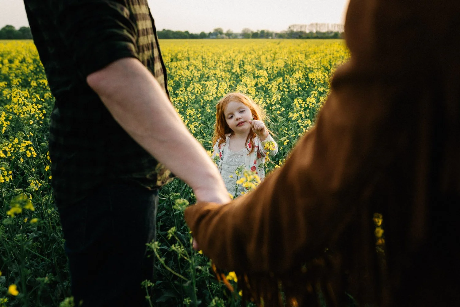 Ein kleines Mädchen mit roten Haaren steht in einem gelben Blütenfeld Rapsfeld, umgeben von zwei Erwachsenen, die sich die Hand halten. Emotionales Familienfoto.
