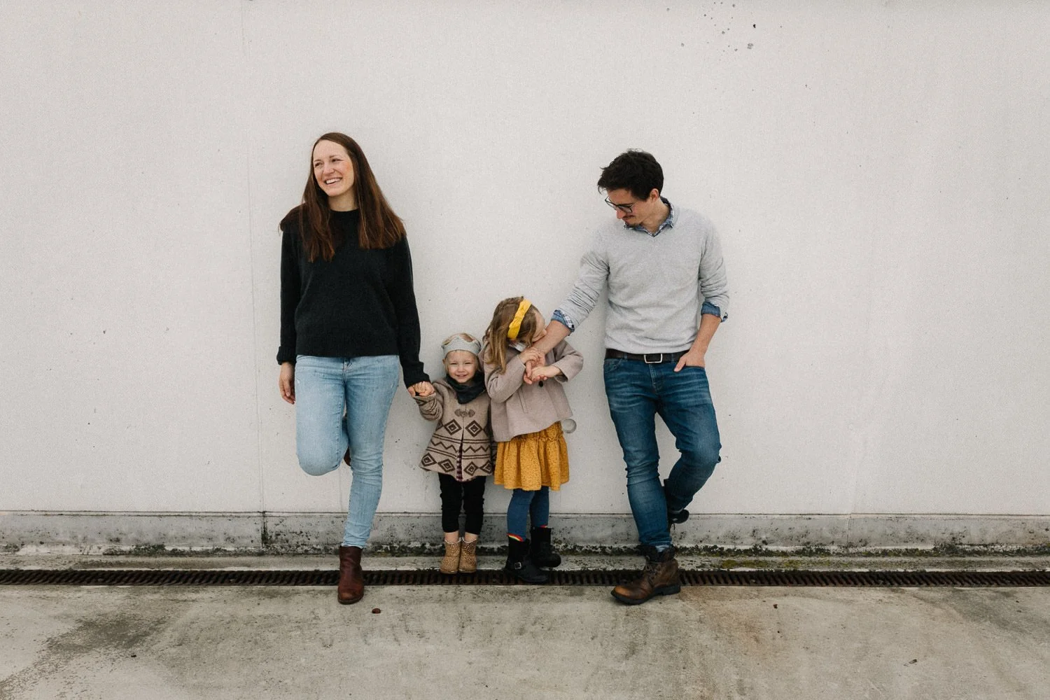Familie bestehend aus zwei Erwachsenen und zwei Kindern, die an einer weißen Wand stehen, lachen und sich gegenseitig an den Händen halten.