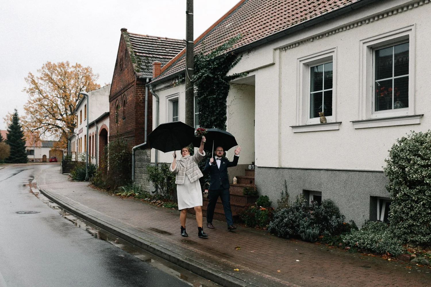 Brautpaar läuft mit Regenschirm Dorfstraße entlang, winken in Kamera