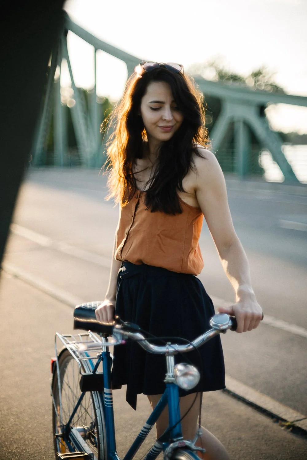 Eine junge Frau mit dunklen, lockigen Haaren, in einem braunen Top und einem schwarzen Rock, steht mit einem blauen Fahrrad auf einer Brücke bei Sonnenuntergang.