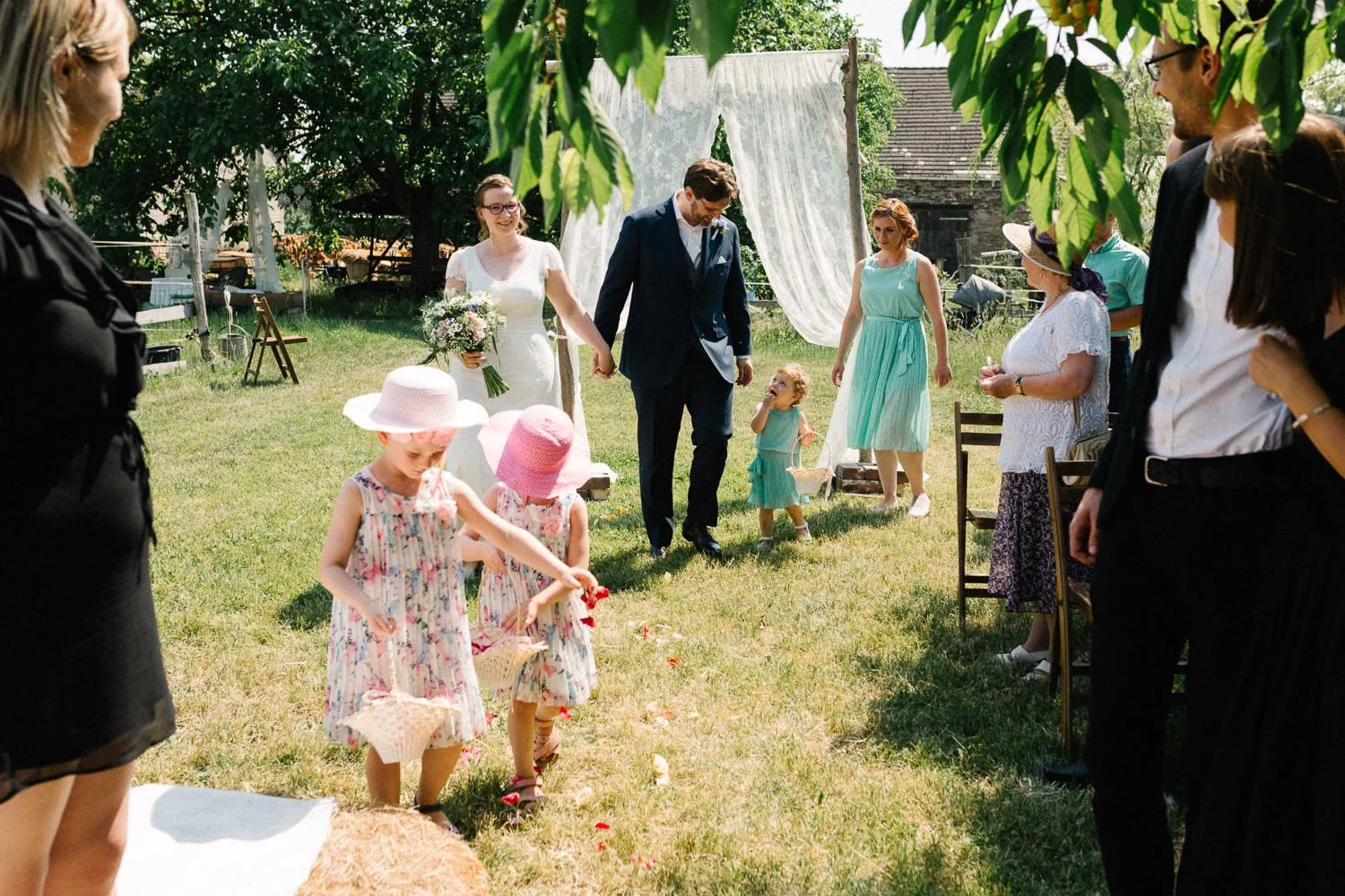 Brautpaar läuft auf Wiese, Hochzeitsgäste rechts uns links, Blumenmädchen vorn