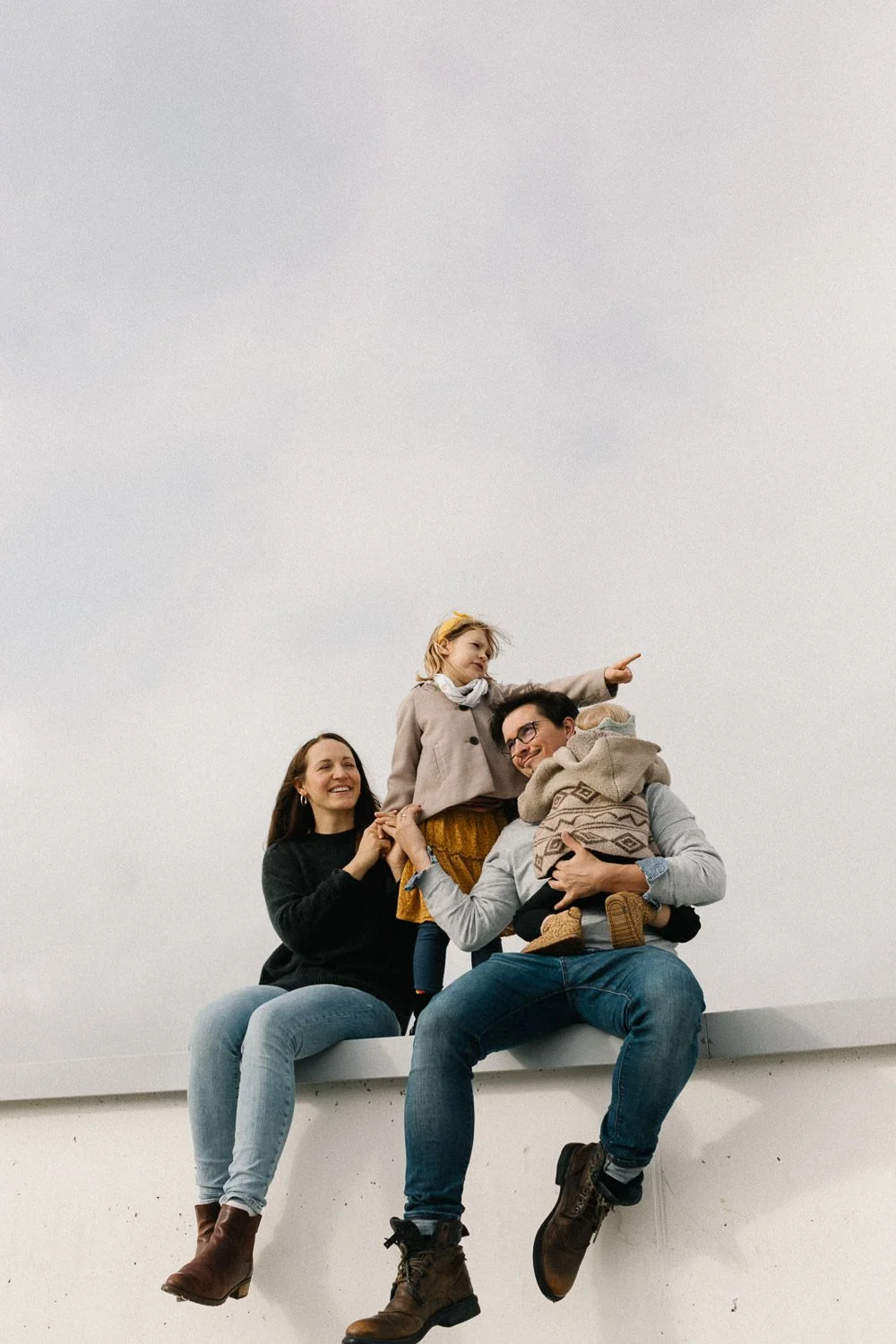 Familie mit zwei Erwachsenen und zwei Kindern sitzt auf einer weißen Mauer im Freien, während das Kind nach oben zeigt. Familienfoto.