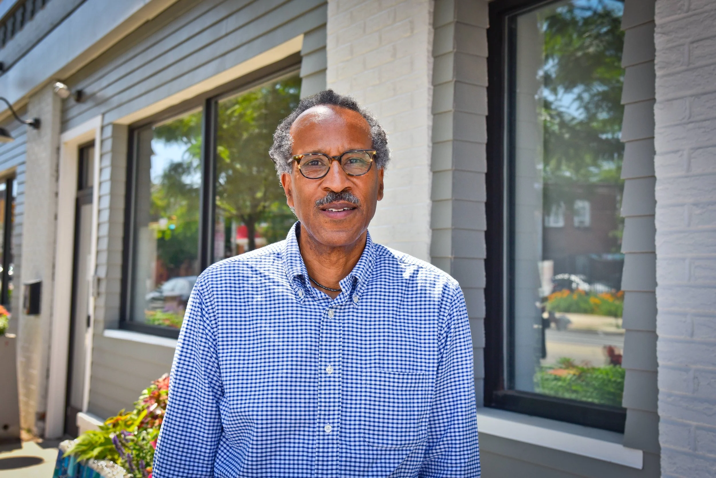 Louis Massiah is standing outside a building with large windows, wearing a blue and white checkered shirt.