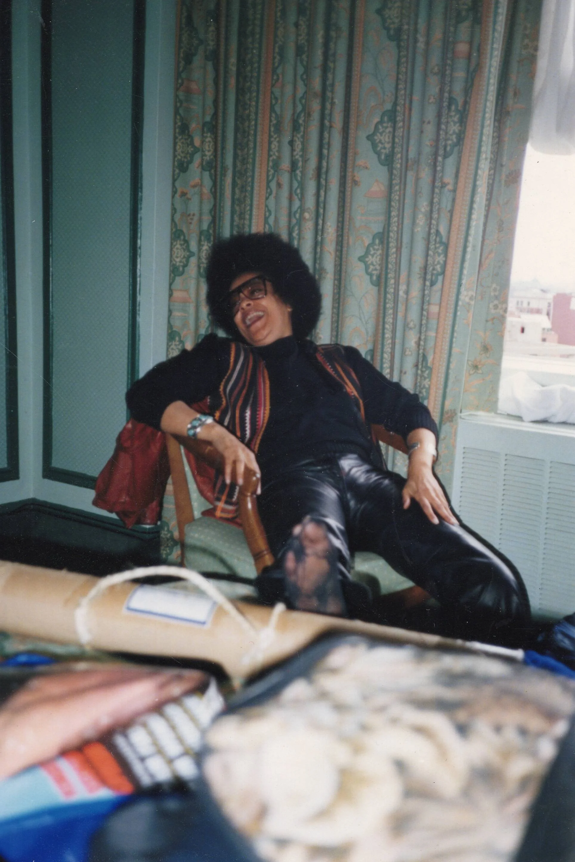 A woman with an Afro hairstyle and glasses, laughing and reclining in a chair in a room with patterned curtains and a window.
