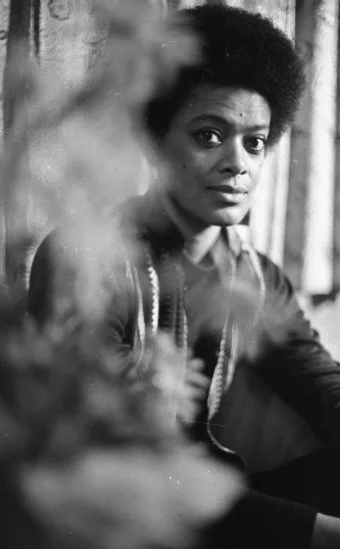 Black and white photo of Toni Cade Bambara, looking directly at the camera, sitting and partly obscured by a blurred object in the foreground.