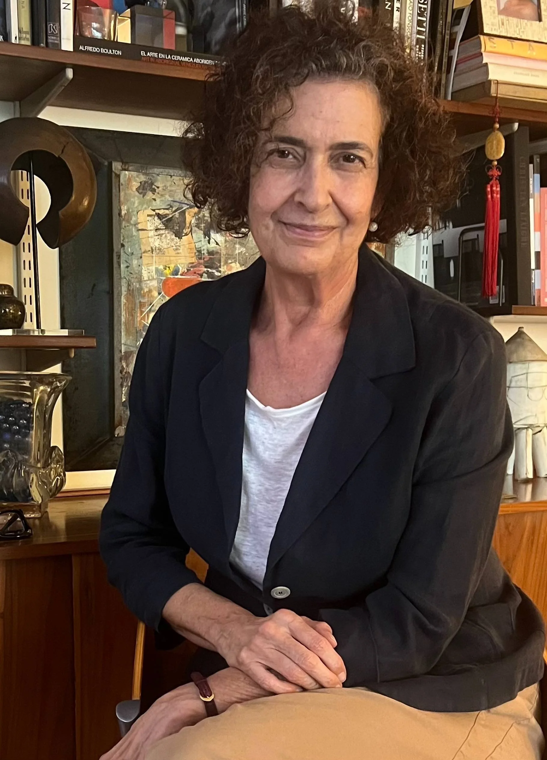 Monica Henriquez wearing a black blazer and white t-shirt, seated in a room with bookshelves and artwork in the background.