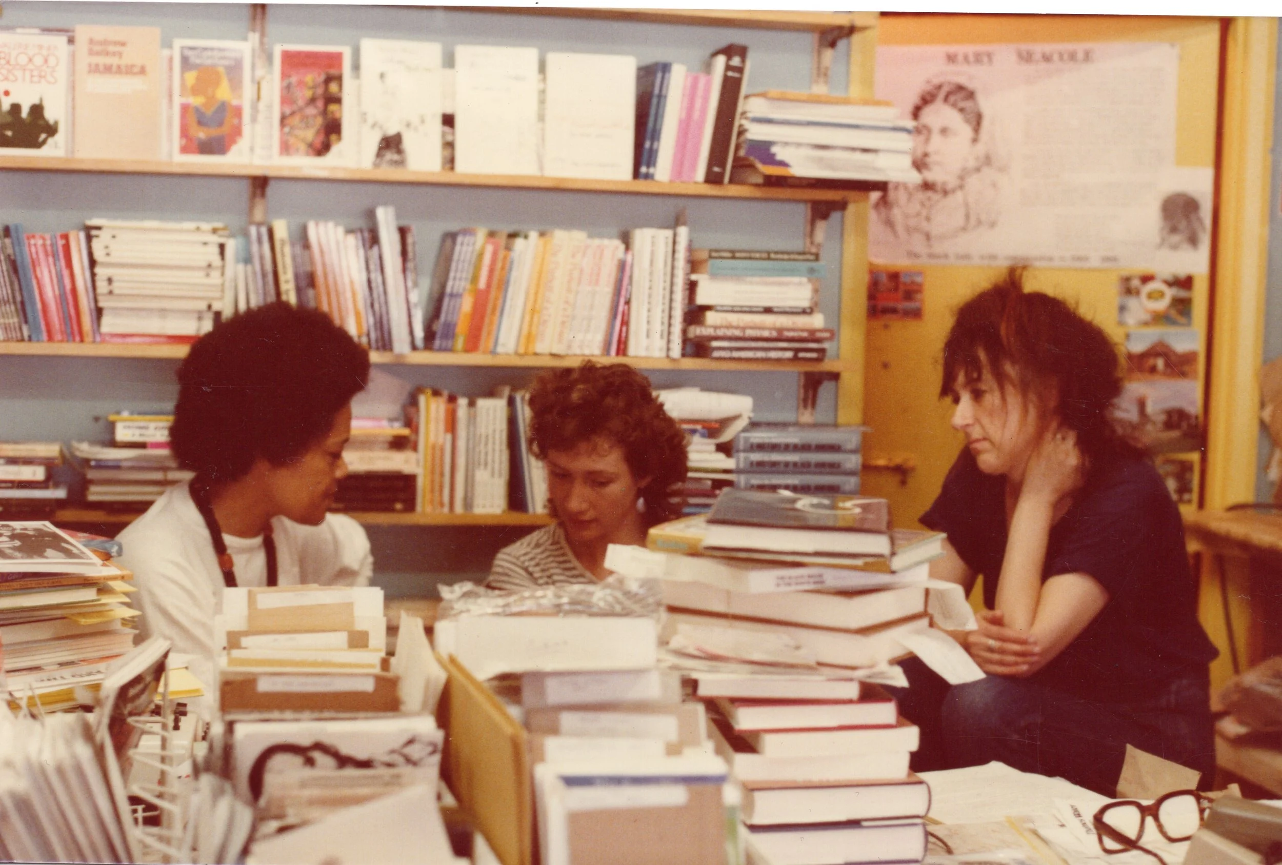 Three women sitting around a table filled with stacks of books in a bookstore or library, with bookshelves in the background.