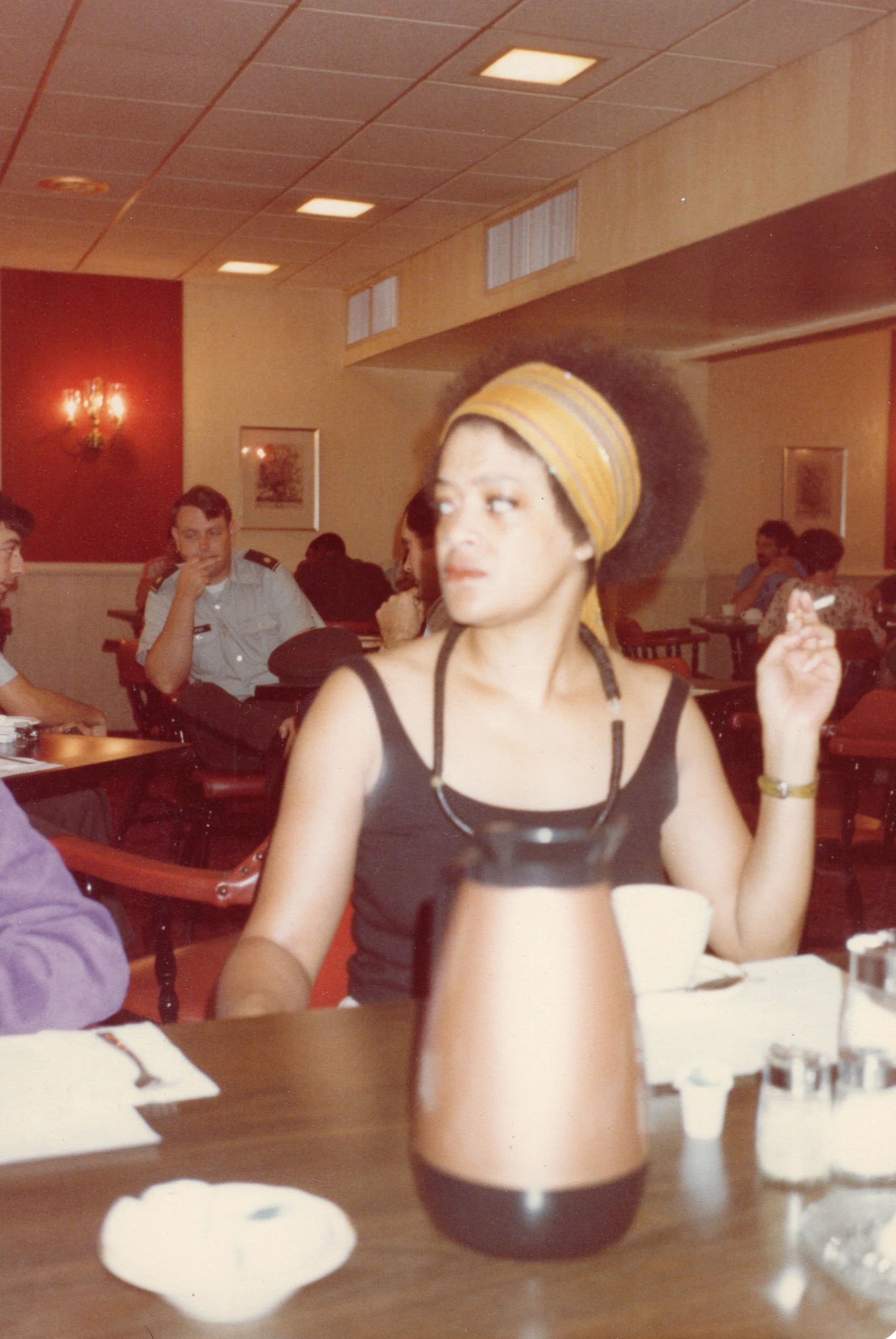 Toni Cade Bambara with a headscarf and dark sleeveless top sitting at a restaurant table and smoking a cigarette