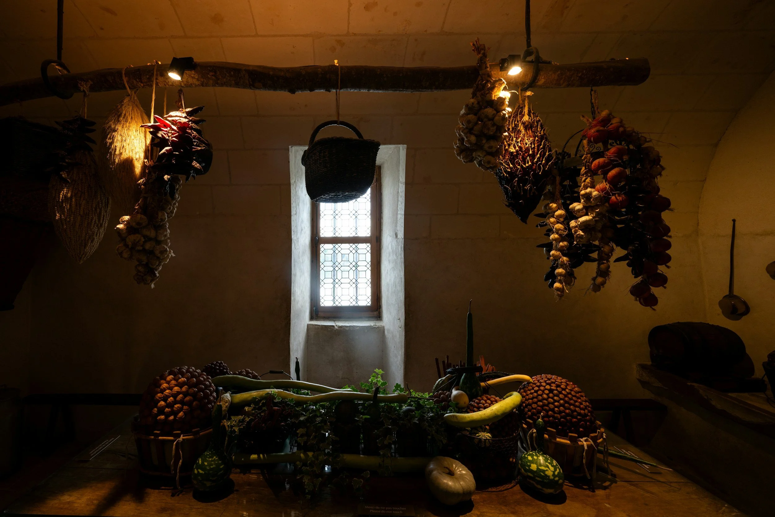 A rustic interior with a small window, hanging dried herbs and garlic, and a table with various vegetables including gourds, squash, and pumpkins.