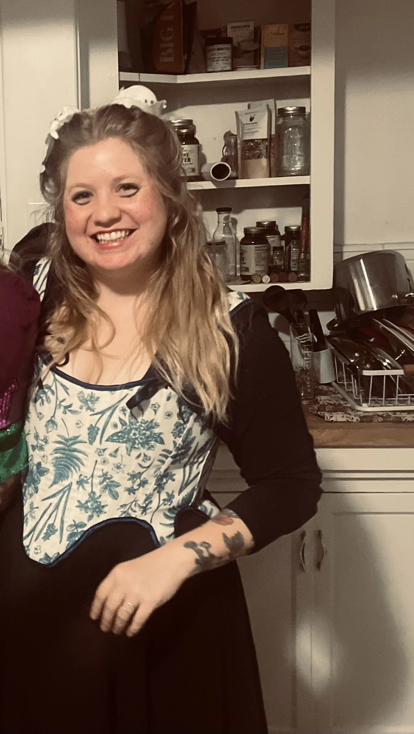 A smiling woman with long wavy blonde hair, wearing a black and white floral apron, standing in a kitchen with open cabinets and a dish drying rack.