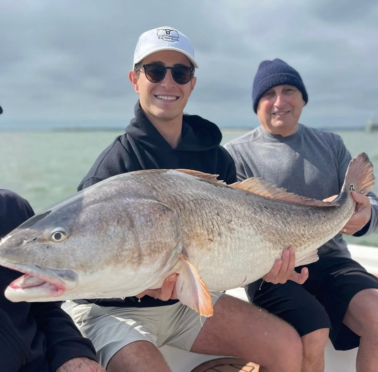 Two men sitting on a boat at the water's edge, holding a large fish together, with one man wearing a white cap and sunglasses and the other wearing a dark beanie, both smiling.