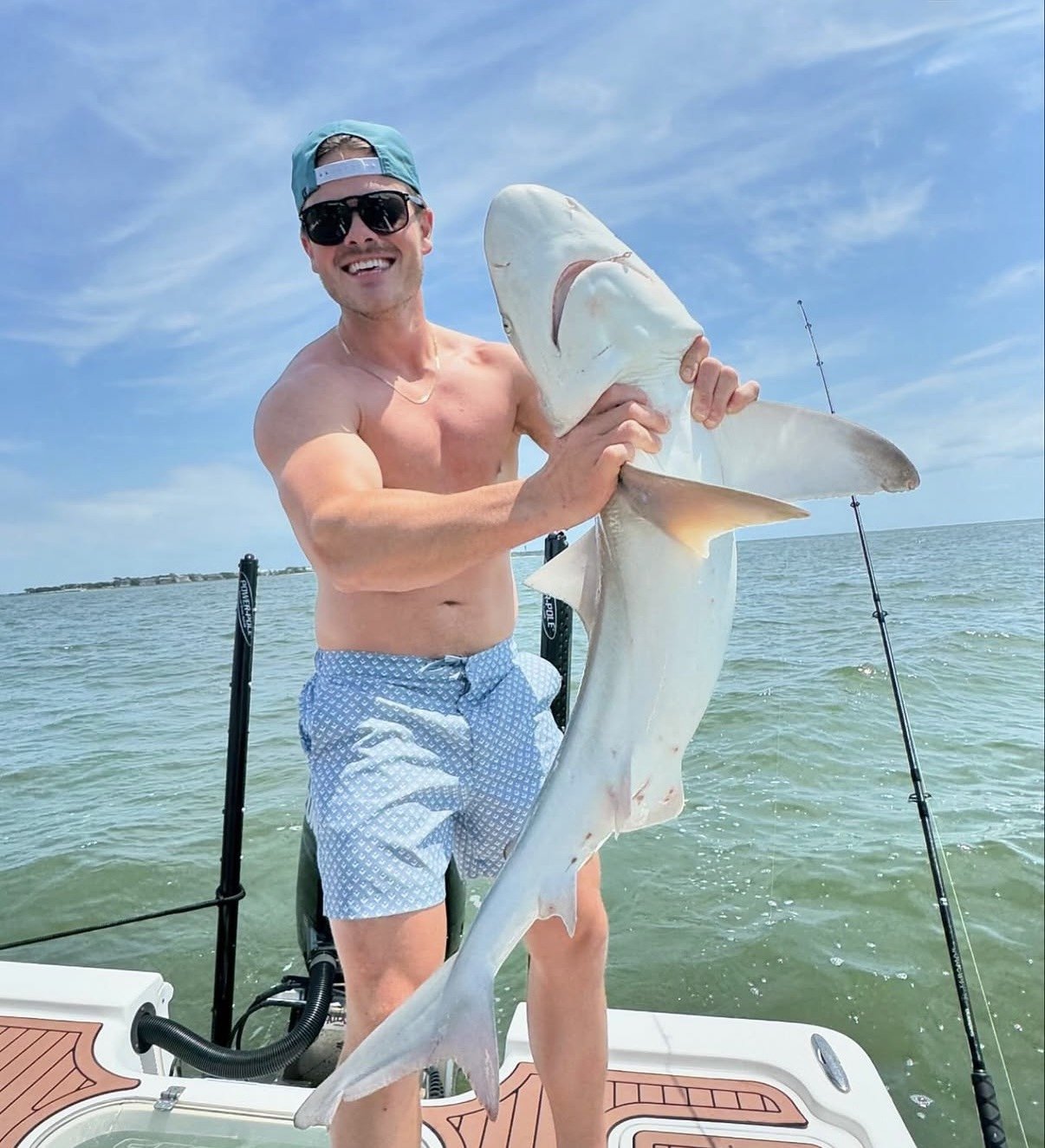 A man wearing sunglasses, a baseball cap backward, and swim trunks, is standing on a boat holding a large shark he caught. The background shows a blue sky, ocean, and fishing rods.