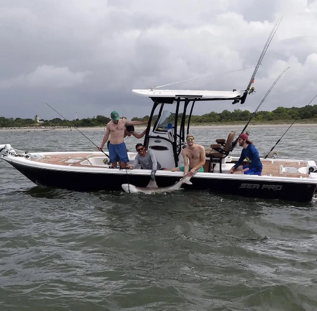 Group of five men on a boat with fishing rods, one holding a large fish. The men are casually dressed, some shirtless, and enjoying fishing on the water. Overcast sky with a shoreline in the background.