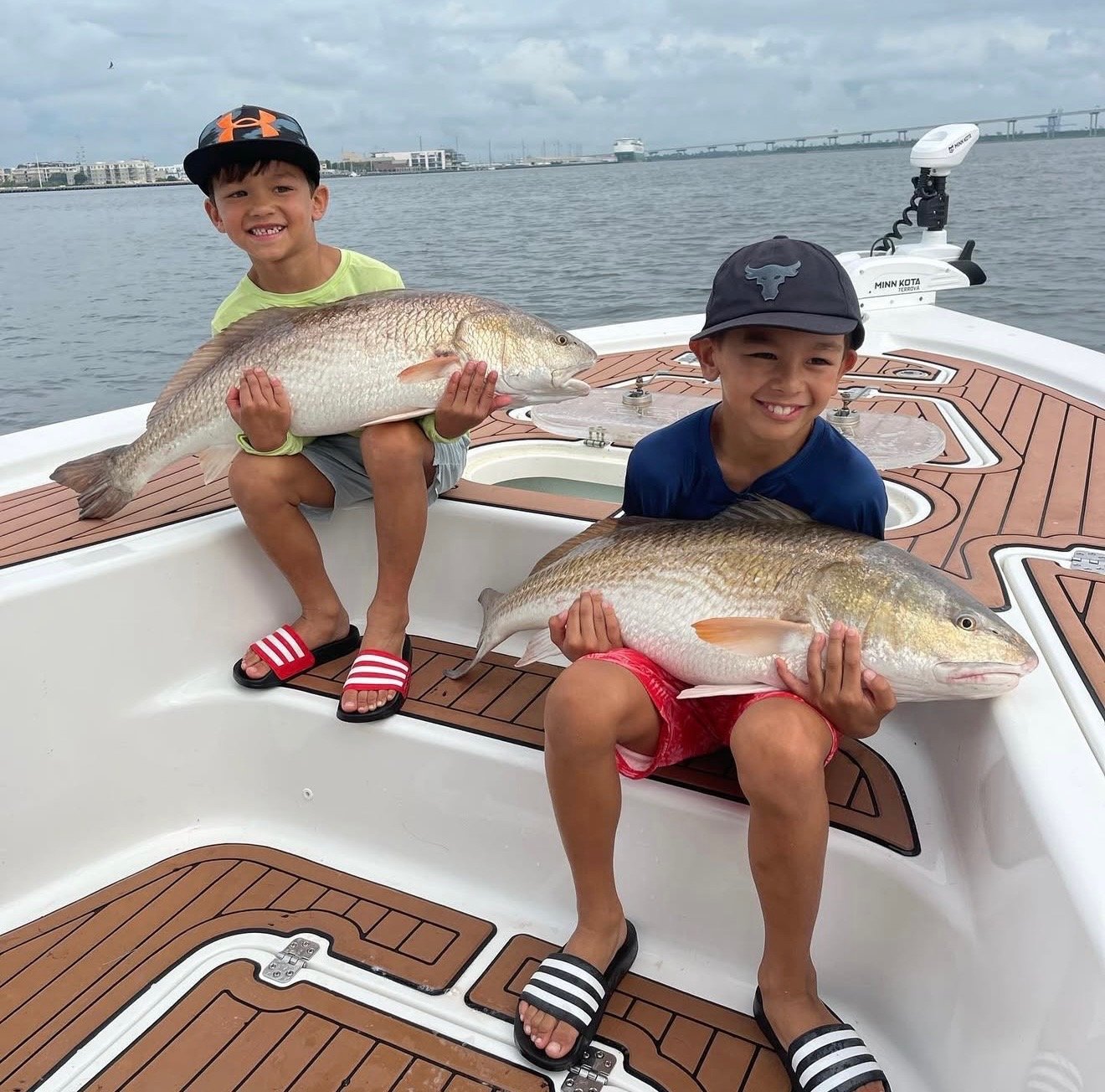 Two young boys sitting on a boat deck, each holding a large fish they caught, with water and a cityscape in the background.