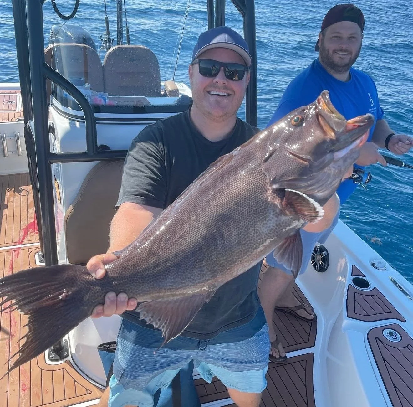 A man in sunglasses holding a large fish on a boat with another person in the background fishing.