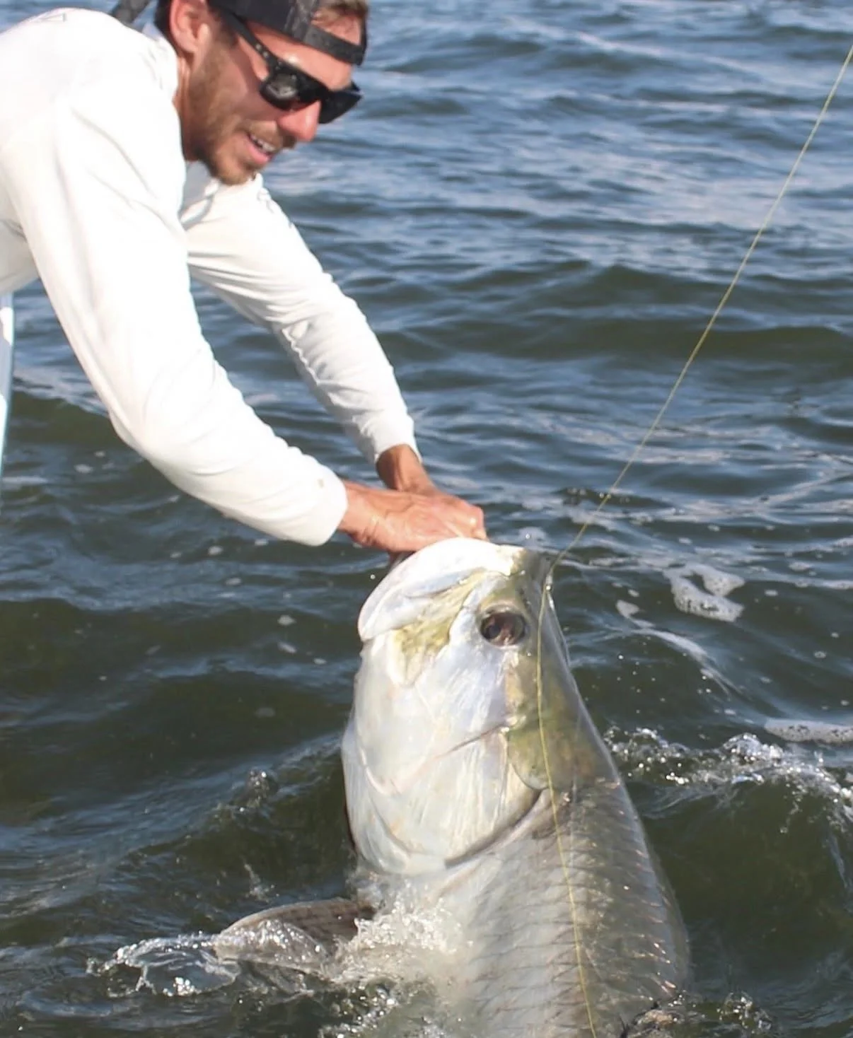 Man in white long sleeve shirt and sunglasses holding a large fish in the water.