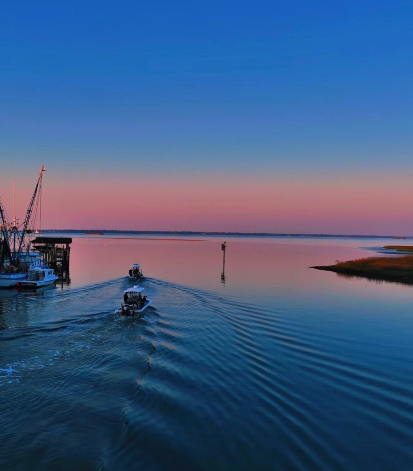 A serene waterfront scene at dusk with small boats on calm water, a pastel sky with pink and purple hues, and a distant shoreline with marshland.