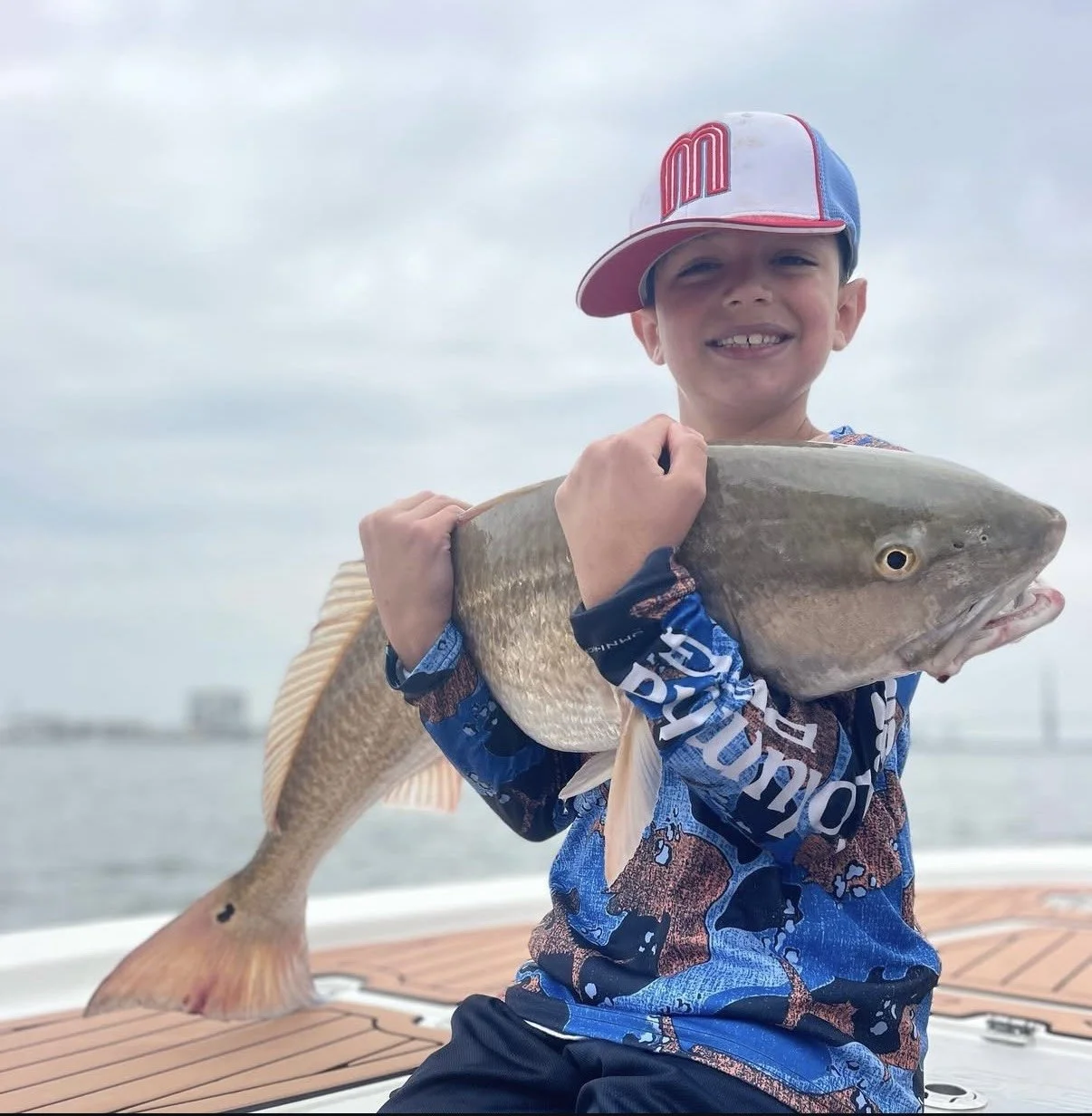 A young boy holding a large fish on a boat, smiling, wearing a baseball cap and a long-sleeve shirt, with a cloudy sky and water in the background.