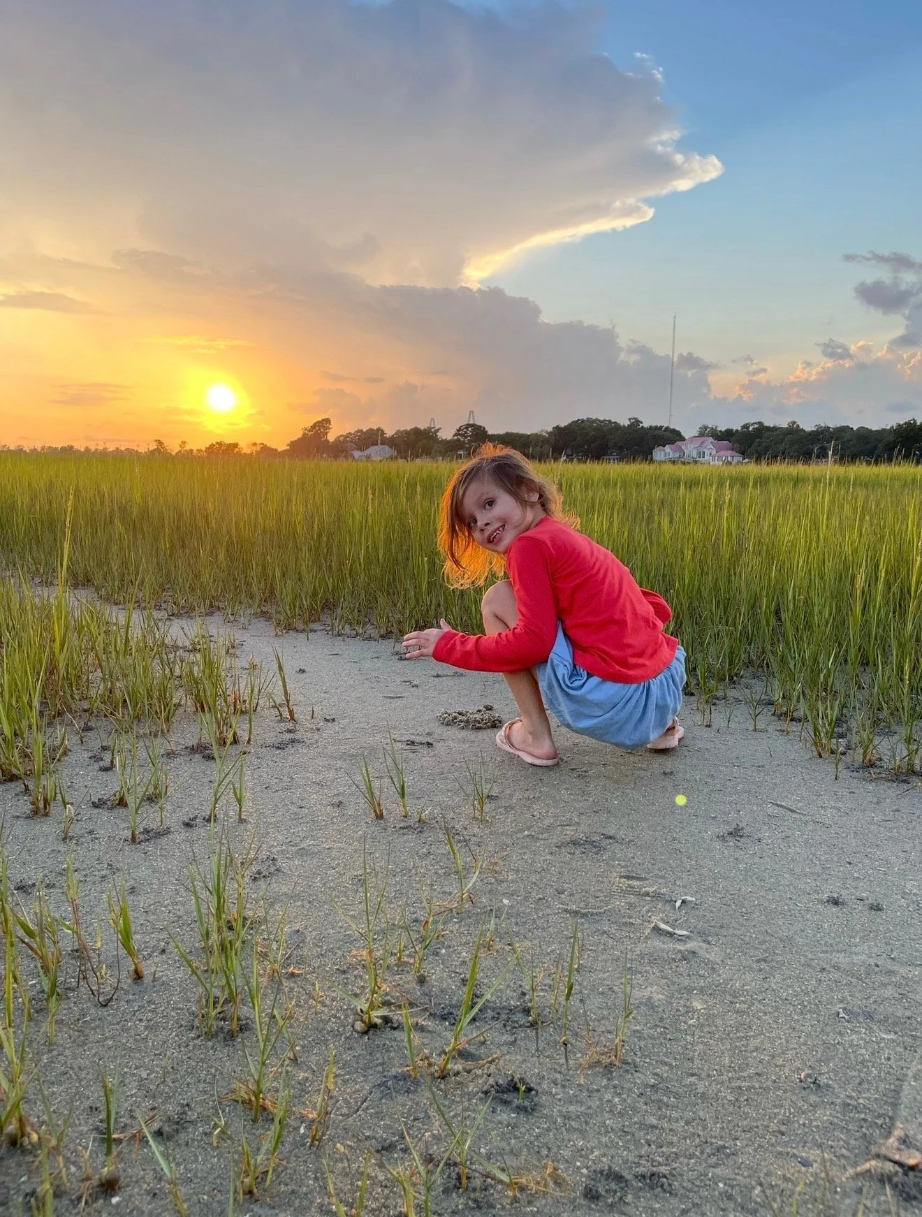 A young girl with brown hair and a pink sweater squatting on a sandy path surrounded by green grass in a field during sunset.