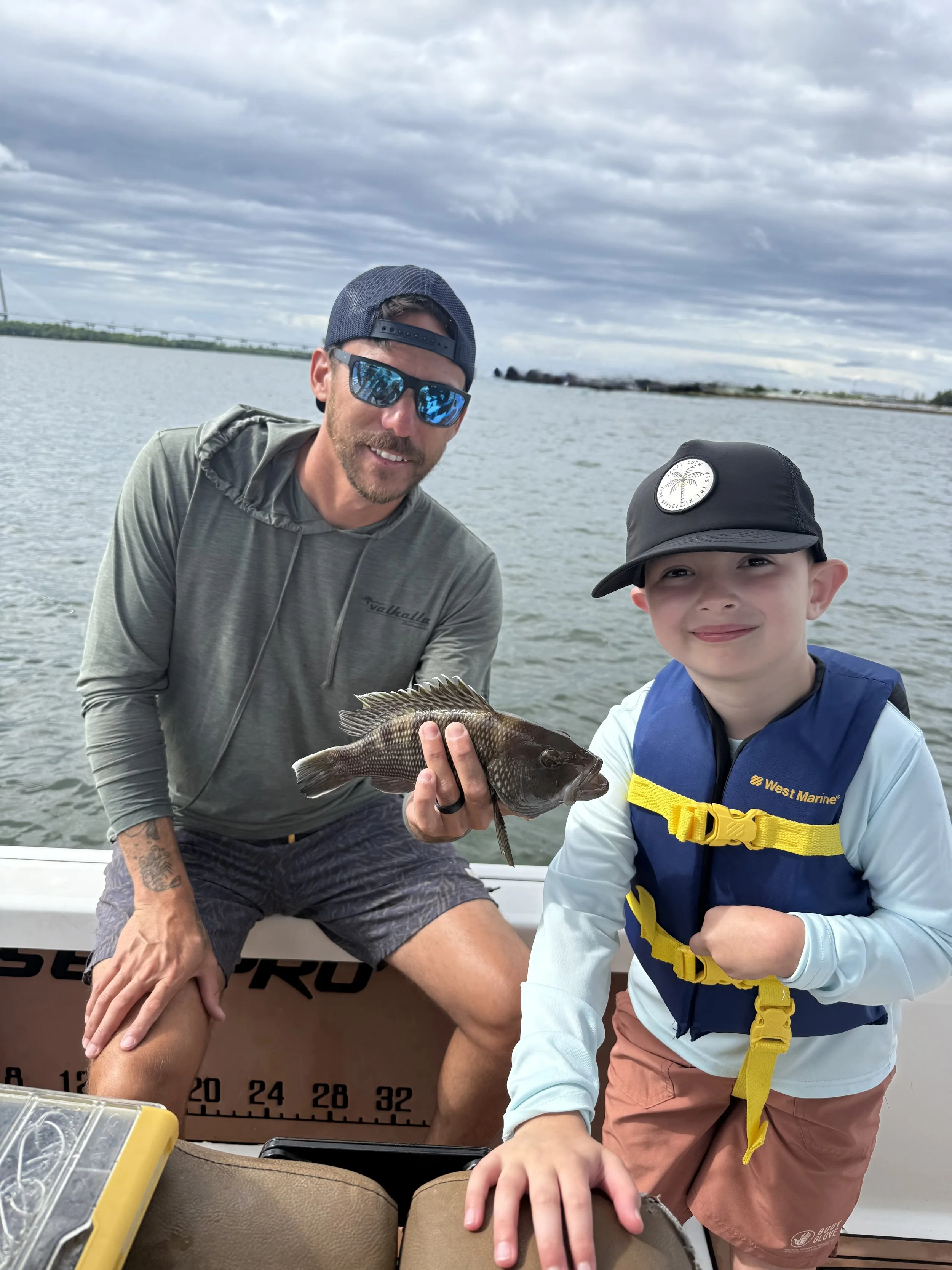 A man and a boy on a boat holding a caught fish, with cloudy sky and water in the background.