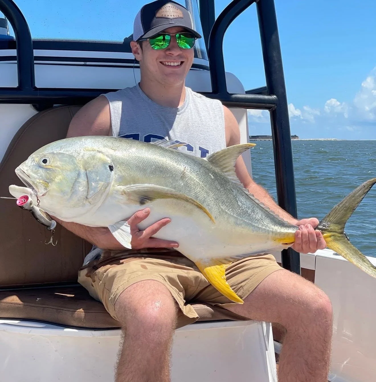 Man sitting on a boat holding a large fish with a yellow tail and a white body, wearing sunglasses, a gray sleeveless shirt, and a baseball cap, during a sunny day on the water.