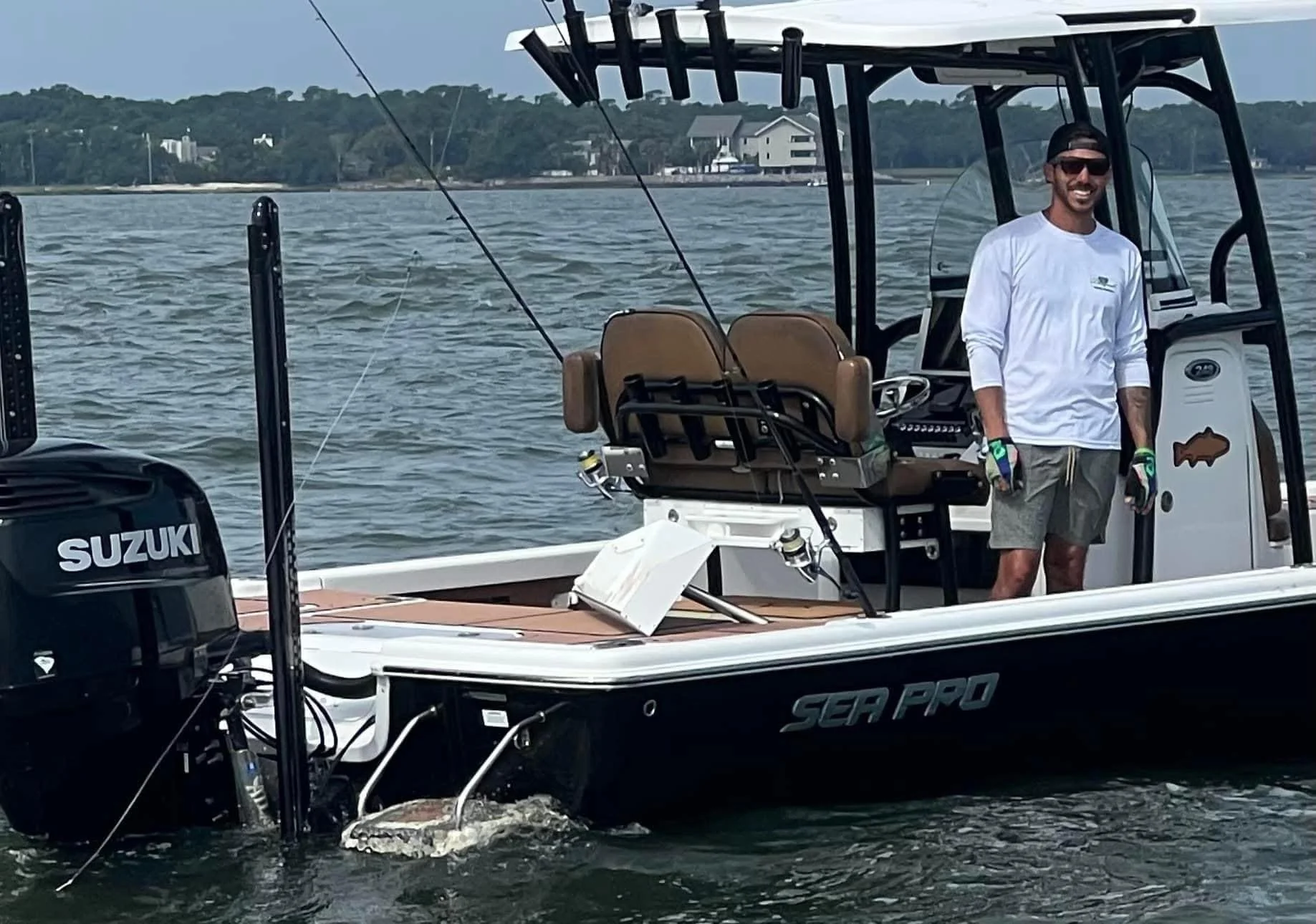 A man standing on a boat near the water's surface, smiling, wearing sunglasses, a white long sleeve shirt, gray shorts, and gloves. The boat has a Suzuki outboard motor and fishing rods attached to it. In the background, there are houses and trees along the shoreline.