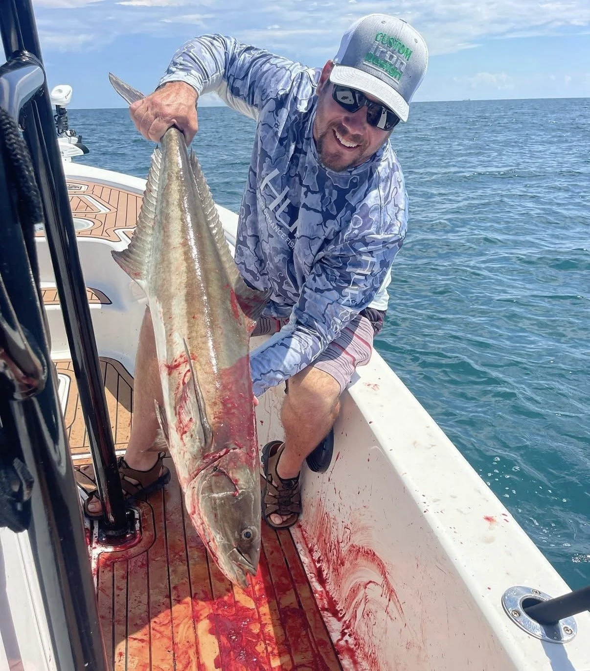 A man on a boat holding a large fish, with blood on the boat's interior, the man smiling and wearing sunglasses, a hat, and casual fishing attire, with an ocean in the background.