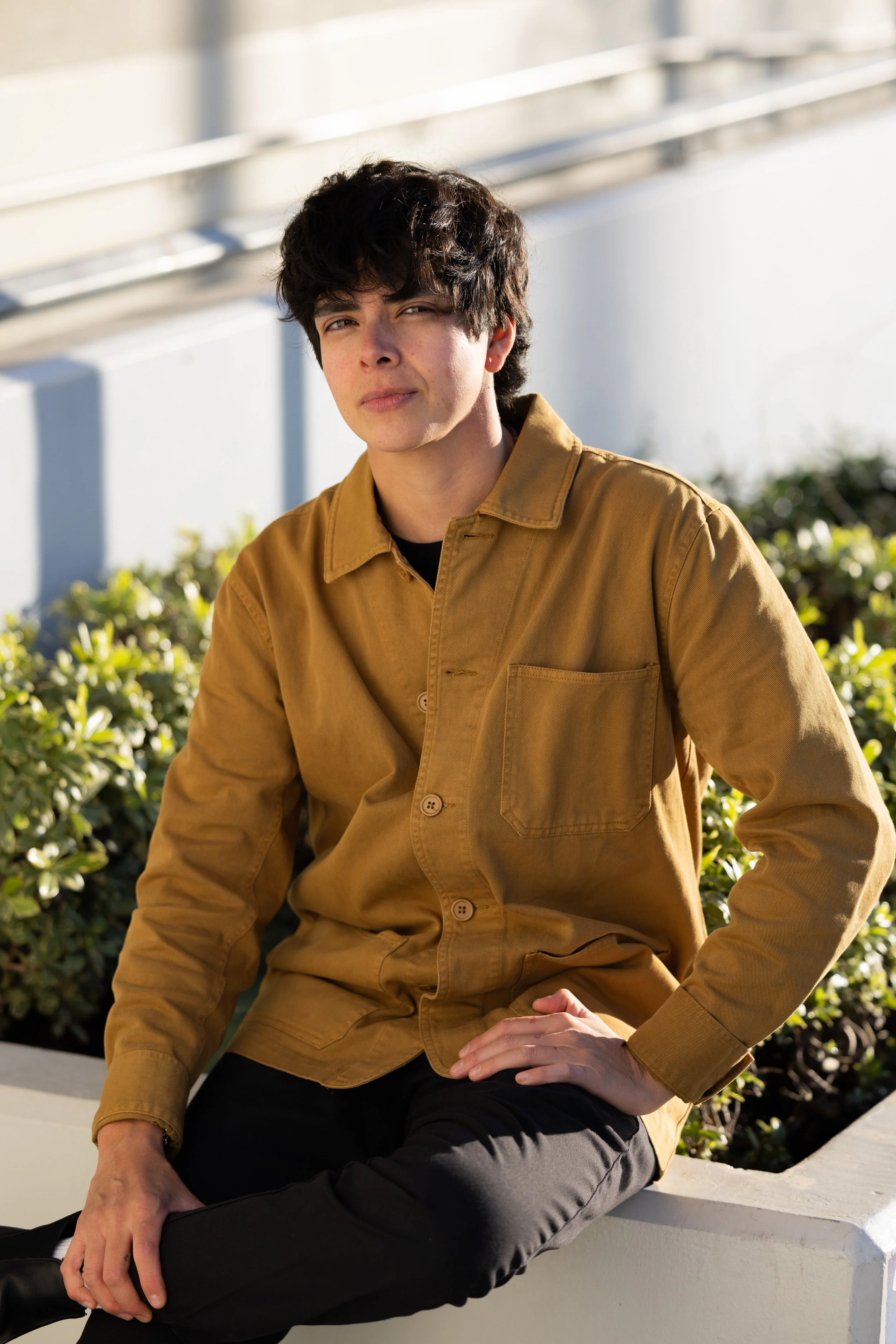 A young man with dark, curly hair sitting outdoors on a white ledge, wearing a tan jacket and black pants, with sunlight on his face and a slightly serious expression. Year After Year Media.  Mini-Documentary.  Non-Profit.  Organization.  Film produc
