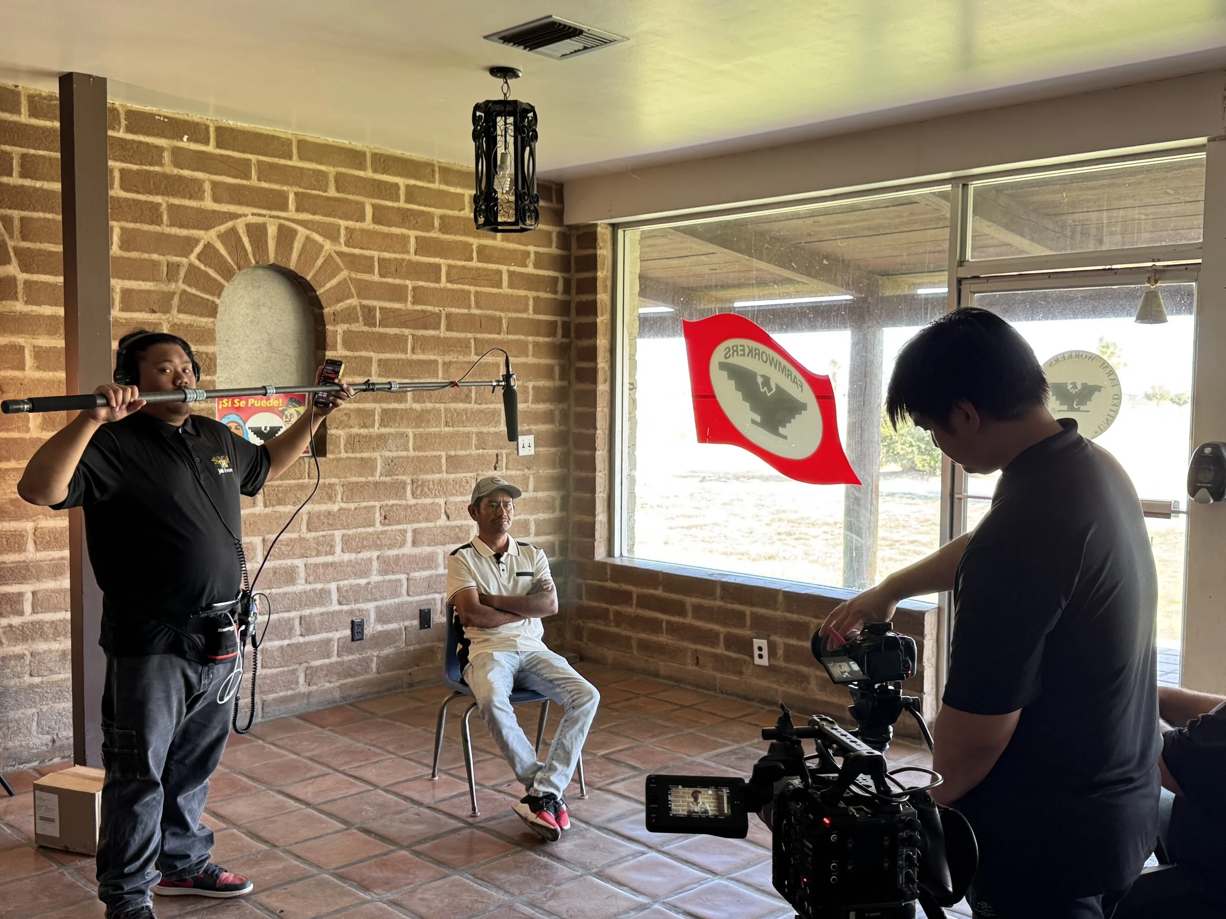 Filming of an interview or documentary with a man sitting on a chair, holding a baseball bat, inside a room with brick walls and a large window. A camera operator and a crew member are present, with the crew member holding a boom microphone.