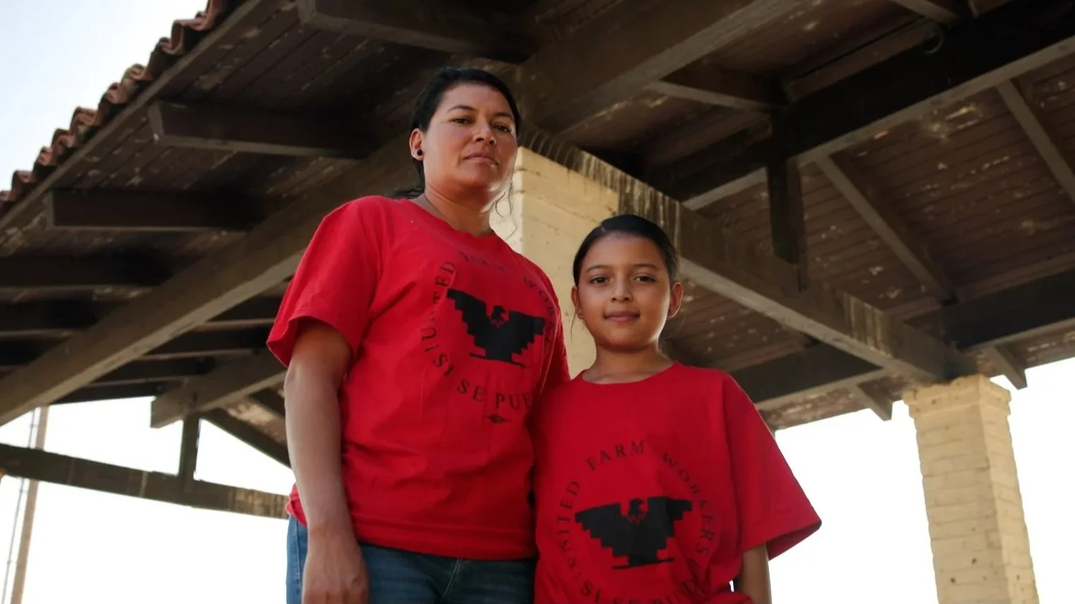 A woman and a girl standing outdoors under a wooden structure, both wearing red T-shirts with a black eagle logo and black text, with a background of a clear sky and brick columns.  Year After Year Media.  Mini-Documentary.  Non-Profit.