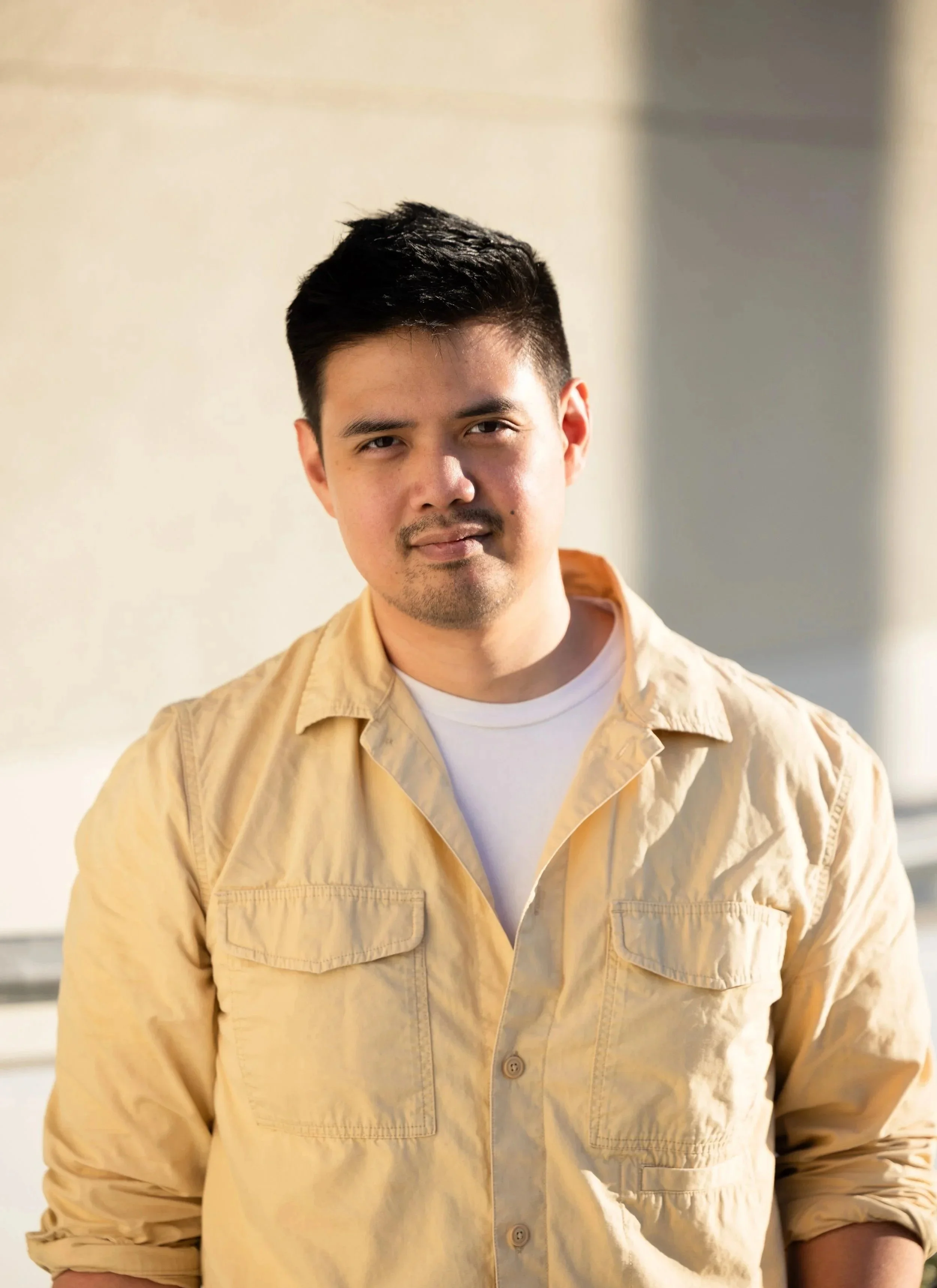 A young man with dark hair and a light beard, wearing a light yellow button-up shirt over a white T-shirt, standing outdoors in sunlight with a neutral expression. Year After Year Media.  Mini-Documentary.  Non-Profit.  Organization.  Film production