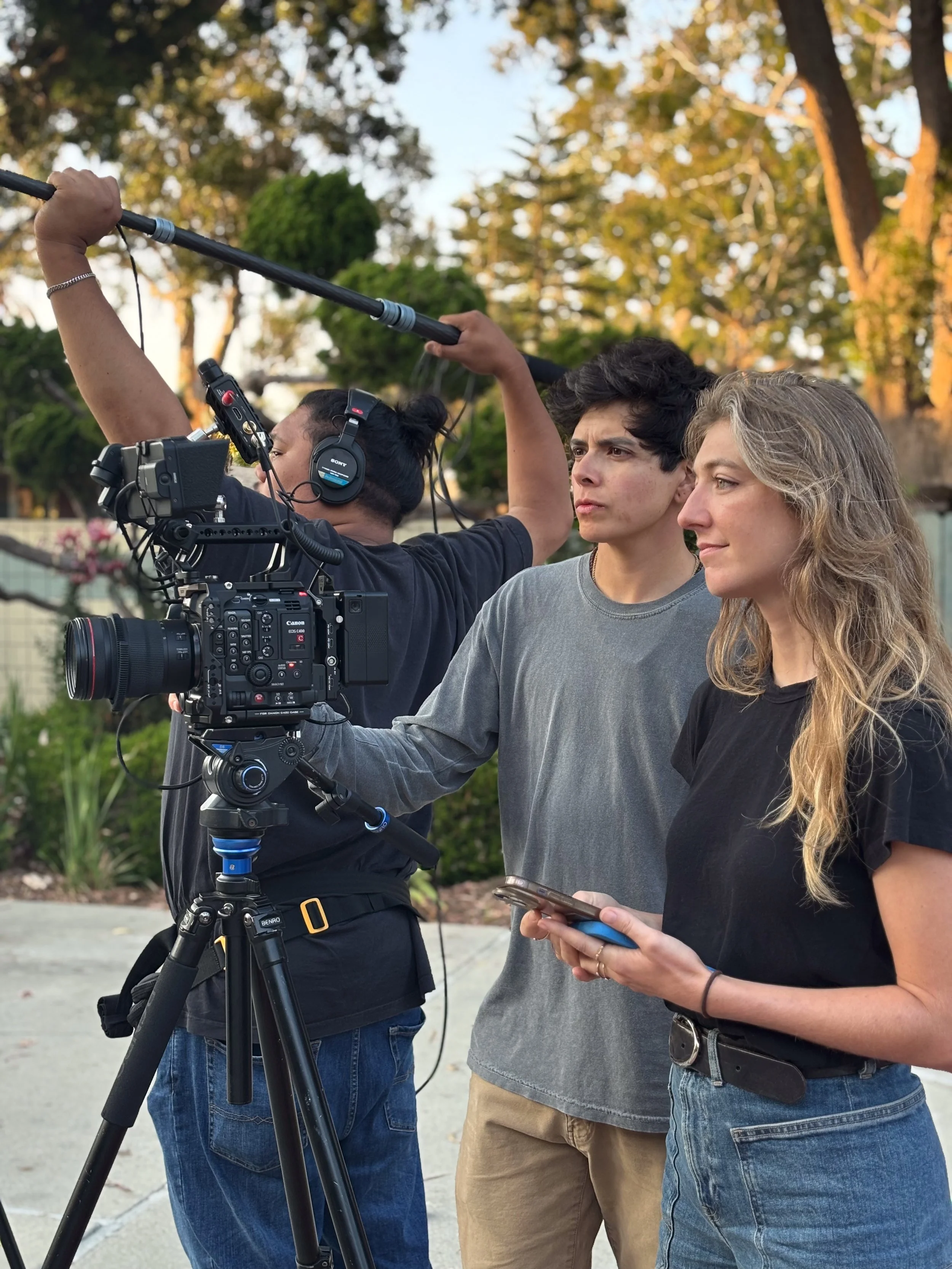 A film crew is filming outdoors during sunset, with a professional camera mounted on a tripod, two crew members operating equipment, and a woman holding a smartphone. Year After Year Media.  Mini-Documentary.  Non-Profit.