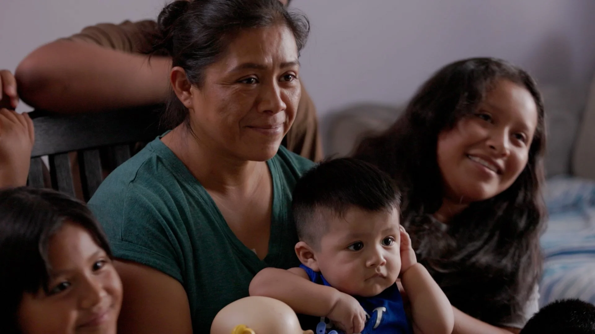 A woman and several children sitting on a couch, with the woman smiling and the children looking in different directions, one girl smiling and a young boy resting his head on her arm.
