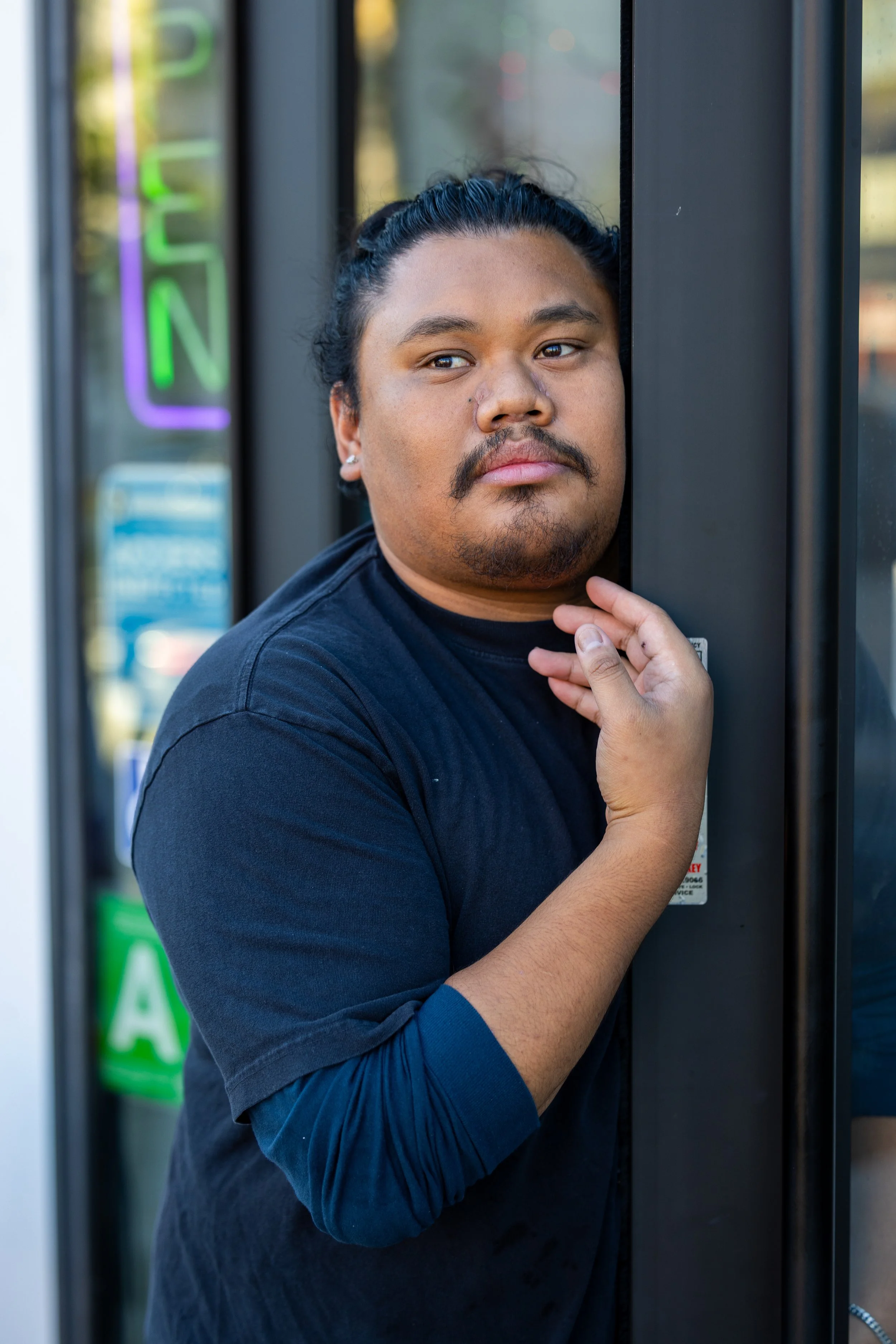A man with dark hair tied back, mustache, and beard stands outside a store, resting his head and hand on the glass door. Year After Year Media.  Mini-Documentary.  Non-Profit.  Organization.  Film production.