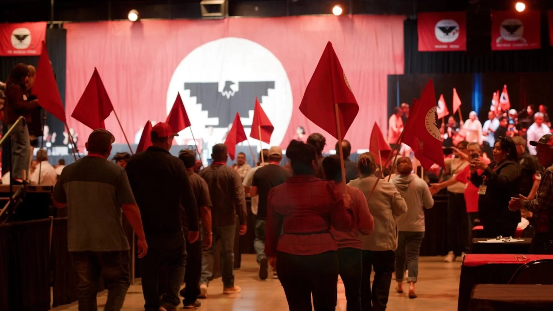 People gathering at an indoor event with flags, a large emblem with wings and a skull on a stage backdrop, and a video screen displaying the same event.