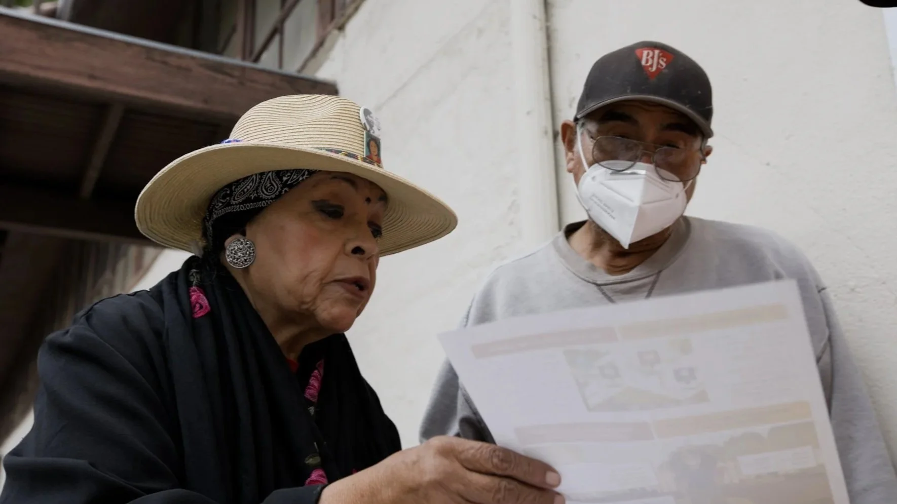 An elderly woman and a man, both wearing face masks, are looking at a document or flyer together. The woman is wearing a wide-brimmed hat, large earrings, and a black scarf, while the man has a baseball cap and glasses.