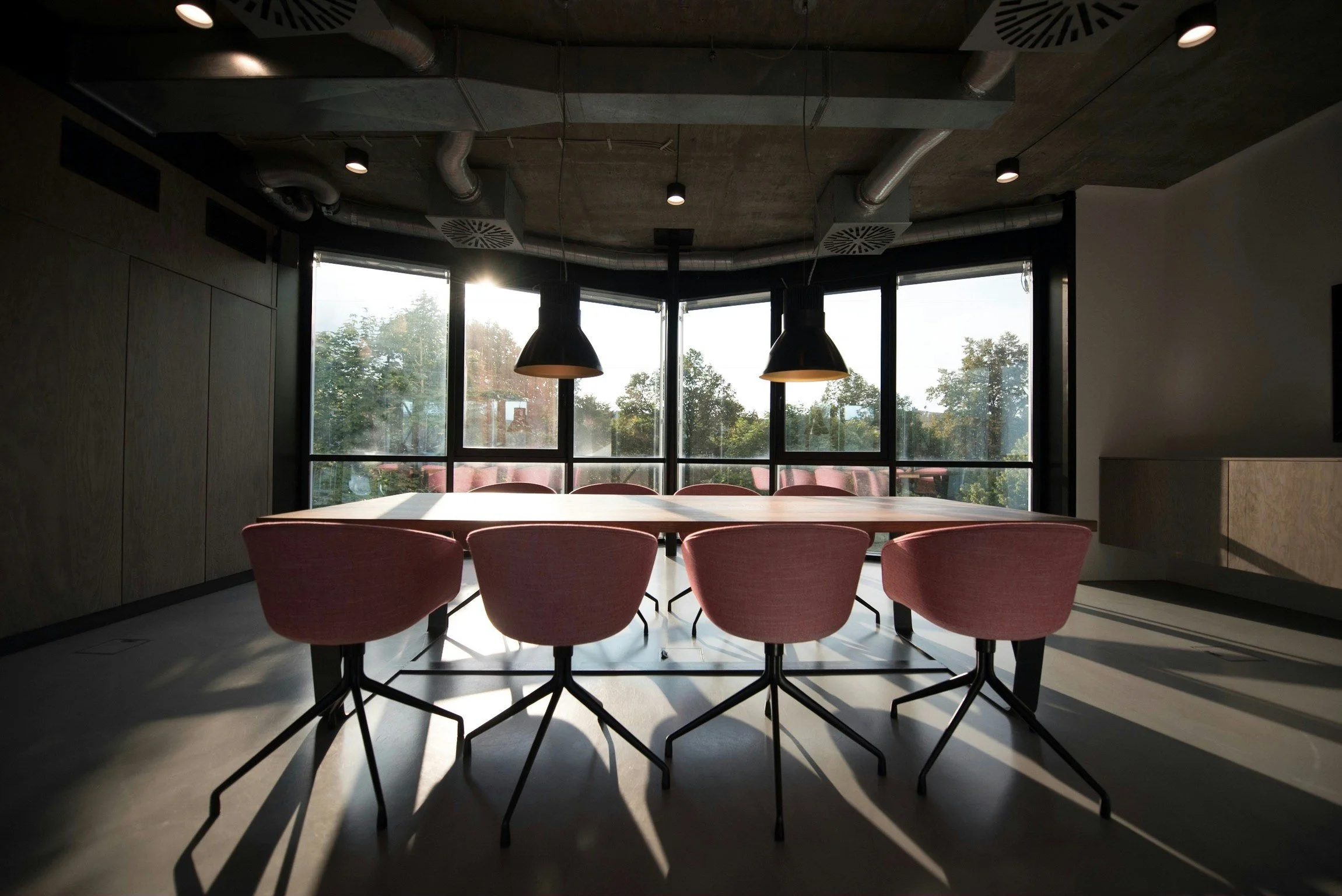 A conference room with a large wooden table, pink chairs around it, black hanging lights, and large windows letting in natural light, with trees visible outside.