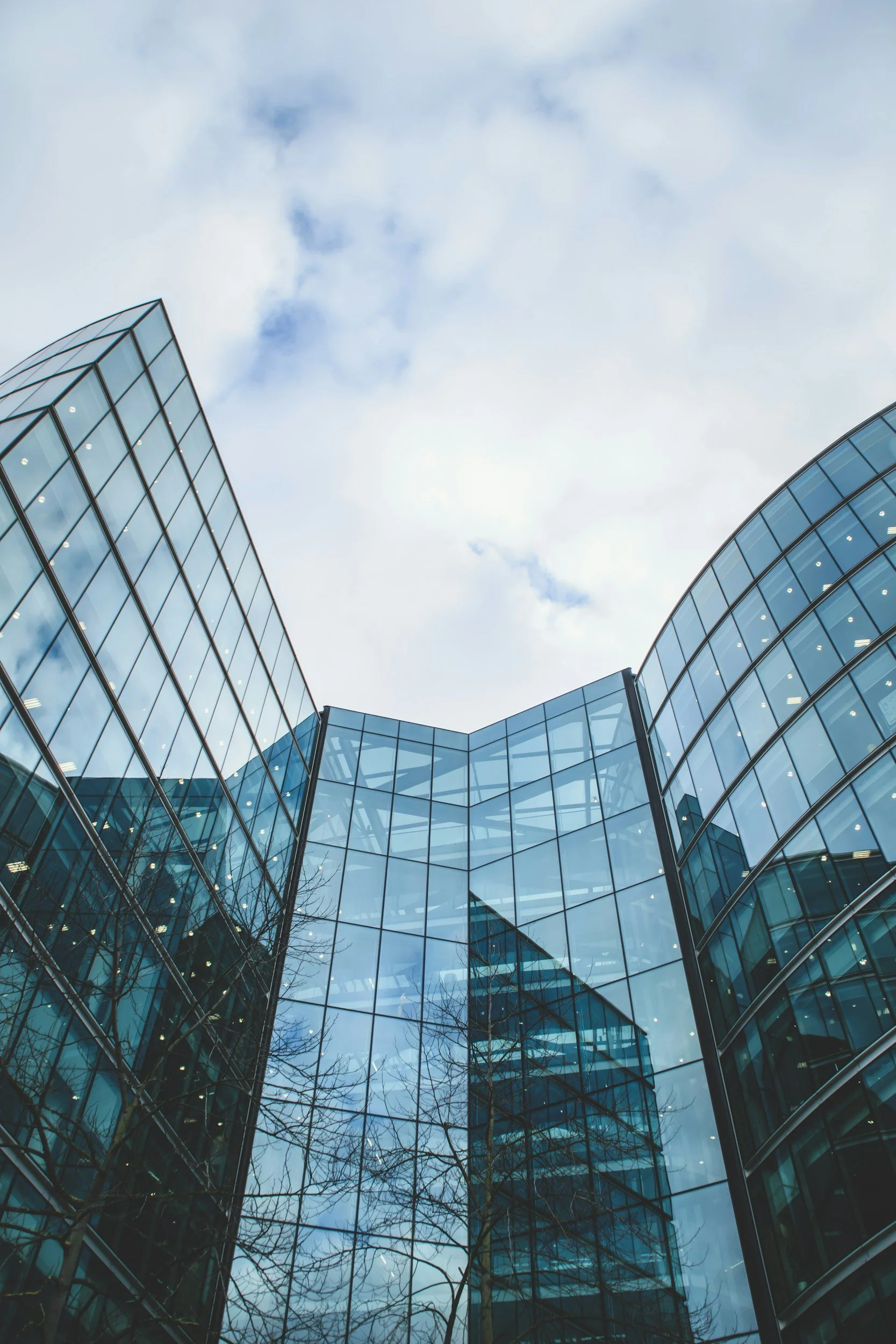 Low-angle view of a modern glass office building reflecting the cloudy sky and leafless trees.