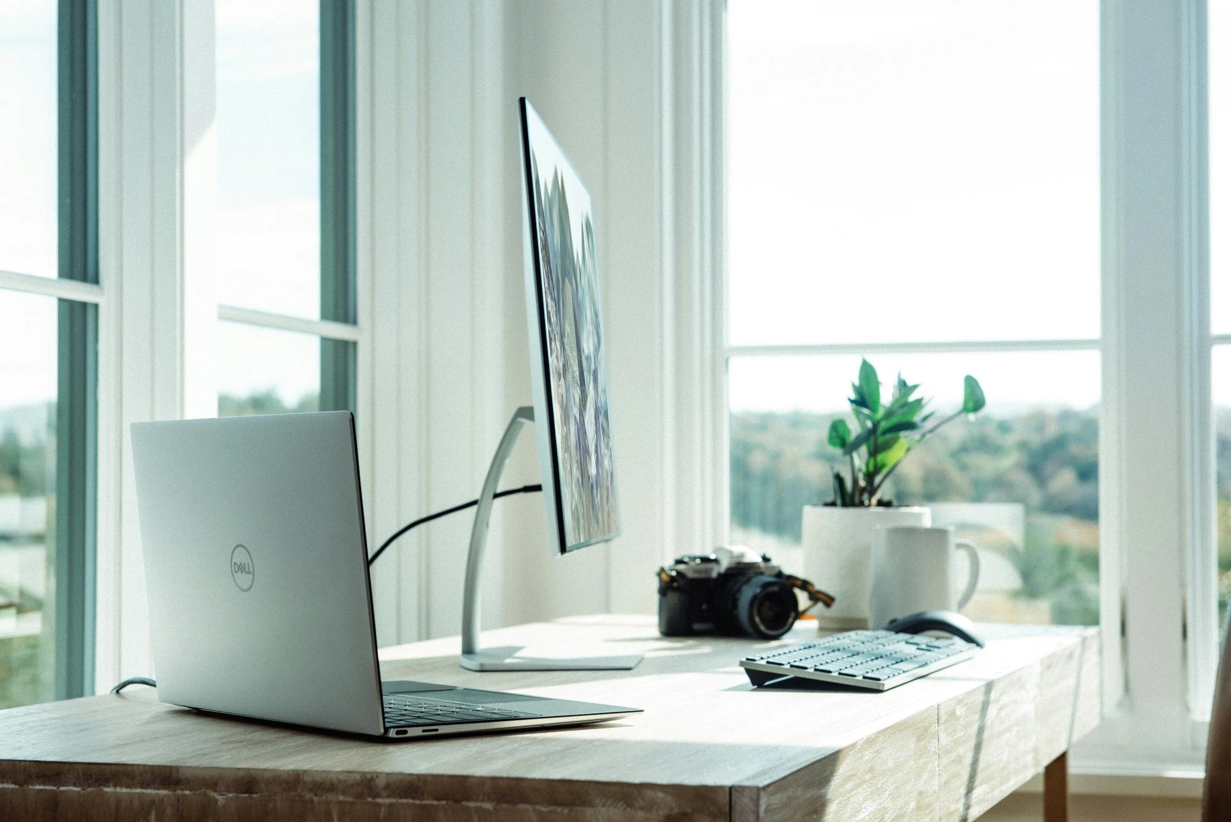 A modern work desk with a silver Dell laptop, a large monitor, a camera, a keyboard, a mouse, and potted plants in front of large windows with a view of the outdoors.