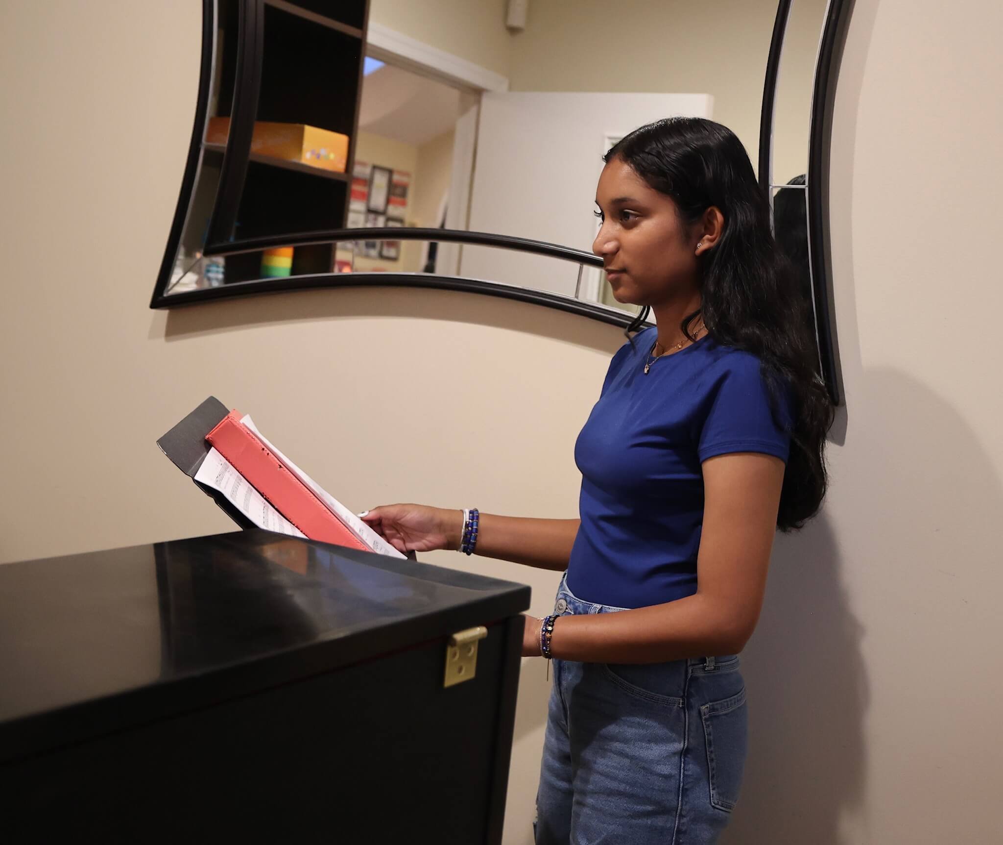 A young woman with dark wavy hair wearing a blue shirt and jeans standing at a black upright piano, holding a pink and white sheet of music, with a mirror on the wall behind her reflecting the scene.
