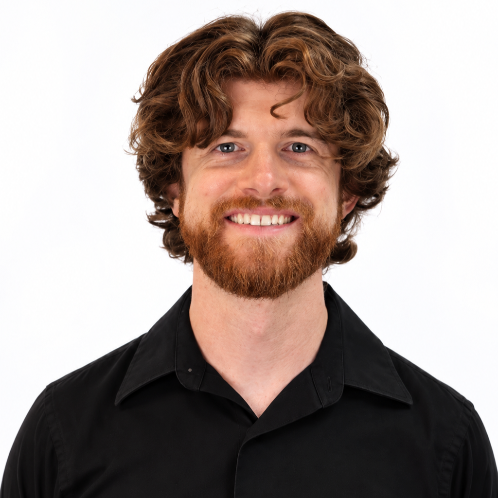 Young man with curly hair and beard smiling outdoors, wearing a light gray sweater over a collared shirt, with trees and greenery in the background.