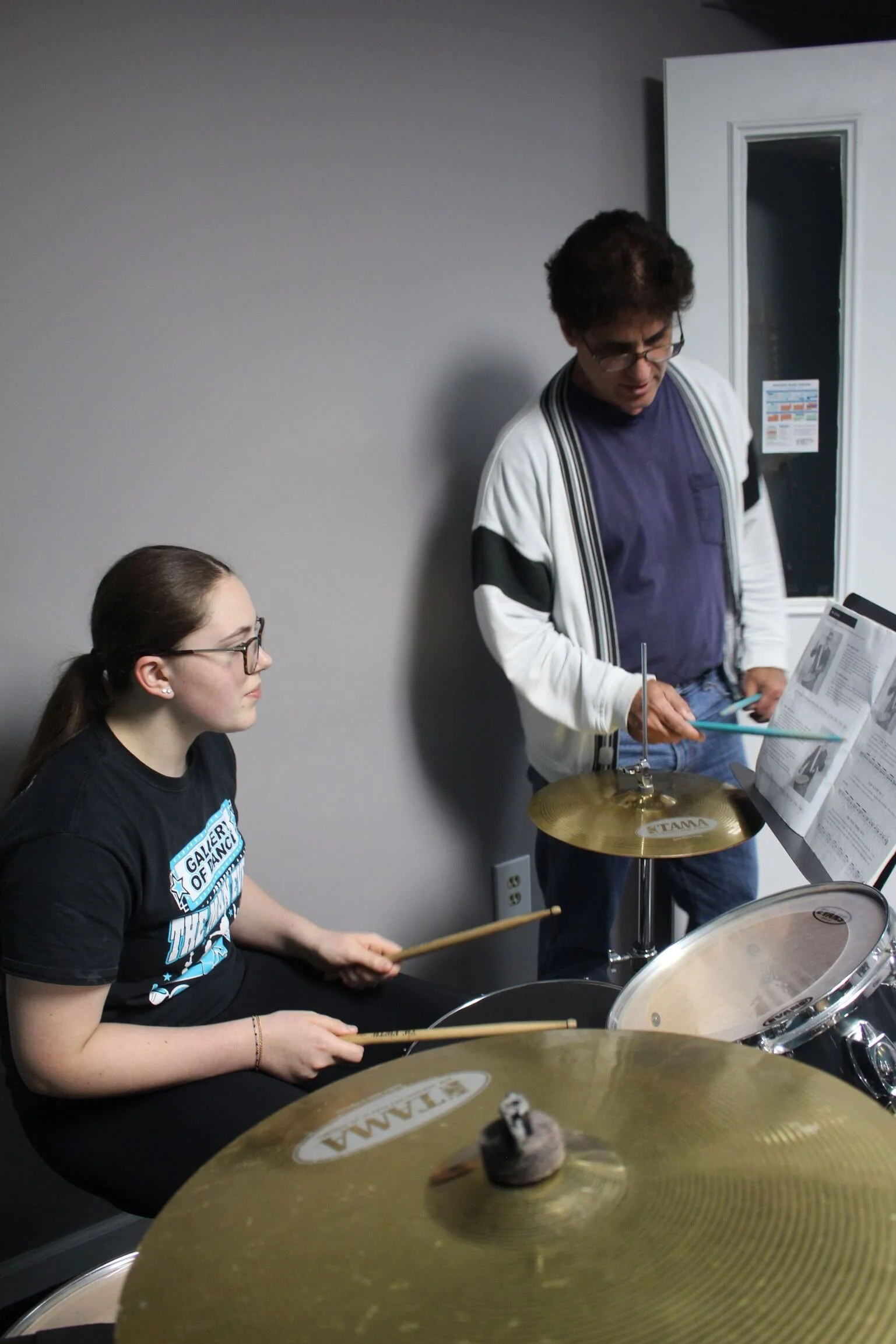 A young woman playing drums and an older man standing next to her, holding drumsticks, in a music room with cymbals and a music stand.
