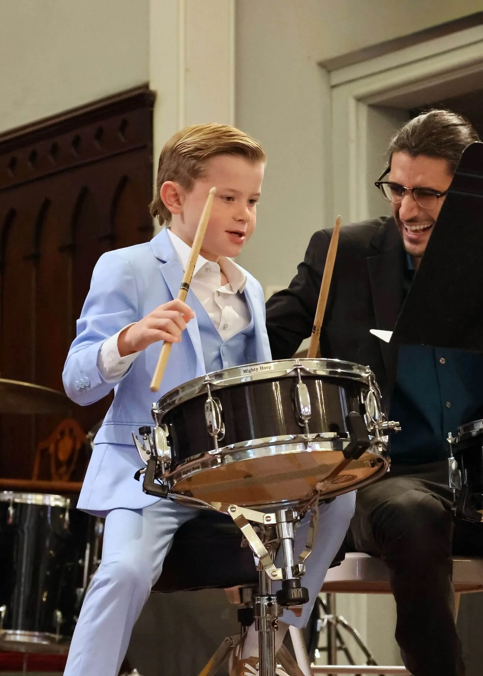 A young boy and an adult man are playing drums together in a room, smiling and enjoying the moment.