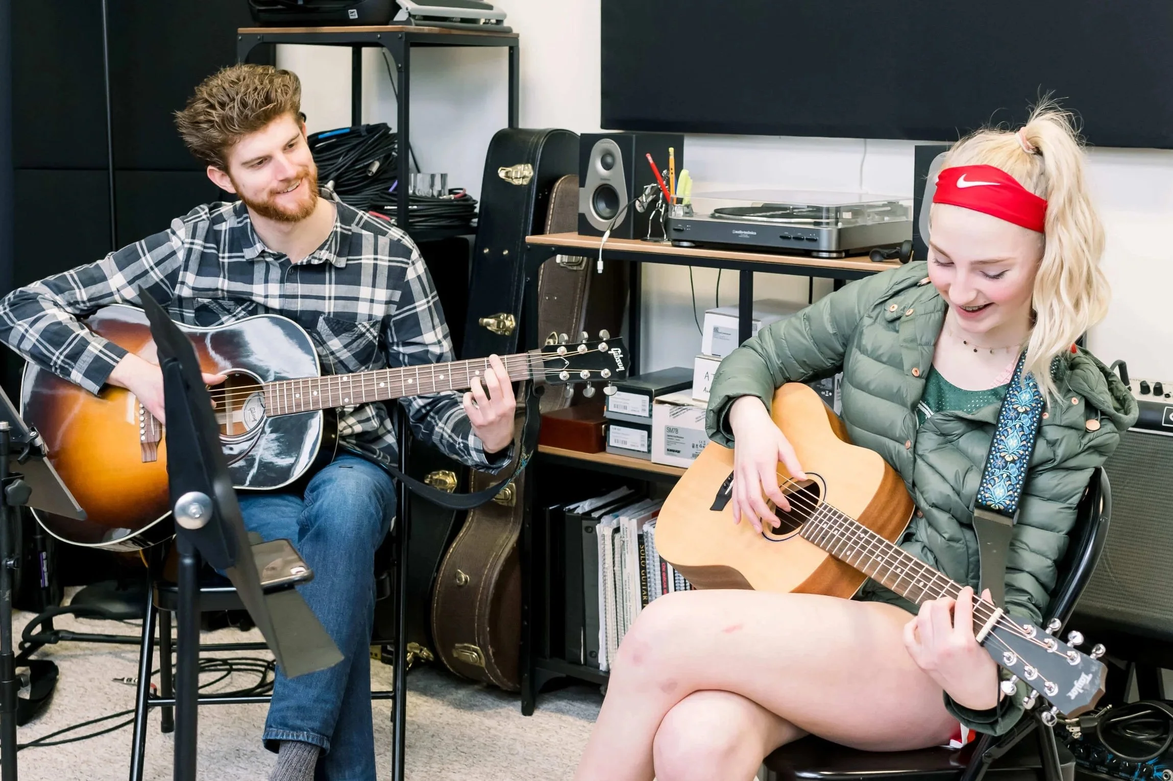 Two young adults, a man and a woman, sitting in a cluttered room, playing acoustic guitars and smiling at each other. The man has curly hair and a beard, wearing a plaid shirt. The woman has blonde hair tied in a ponytail with a red Nike headband, and is wearing a green jacket. There are various electronic equipment, shelves with books, and a turntable in the background.