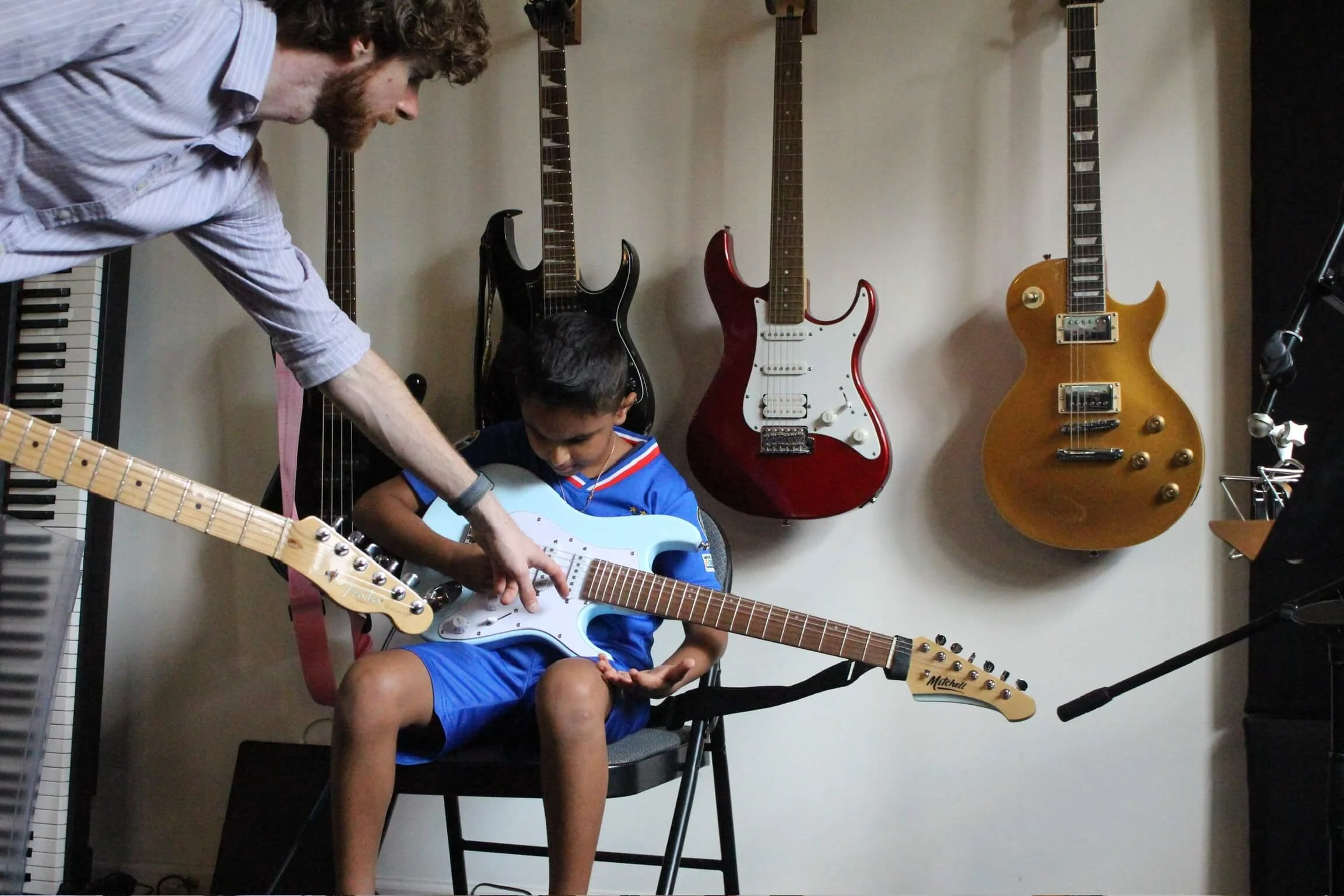 A man helps a young boy learn to play the electric guitar in a music room with several other guitars hanging on the wall and a keyboard on the side.