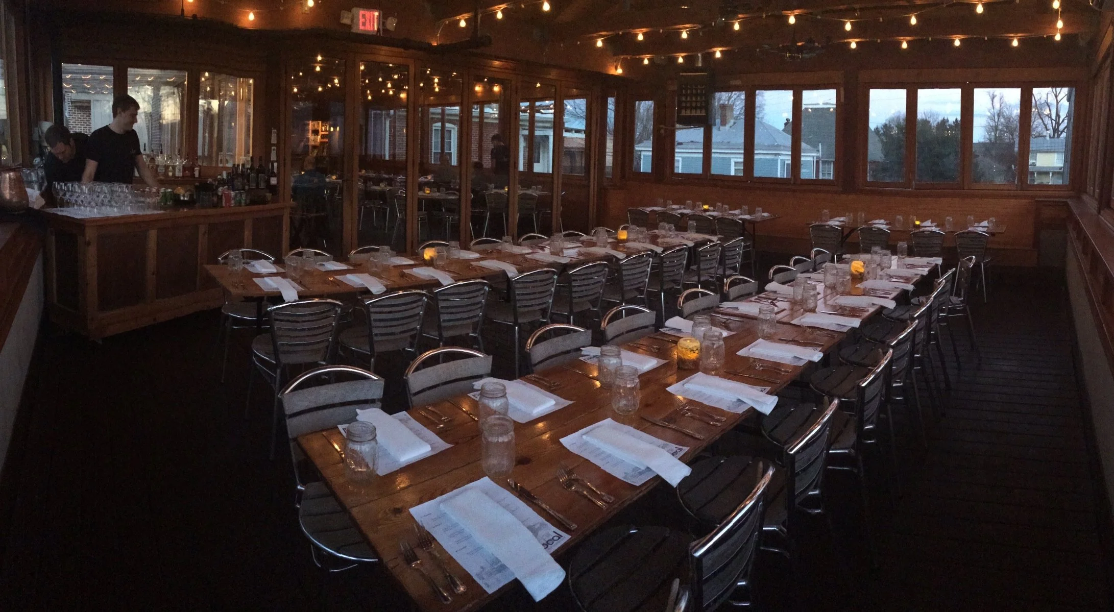 Upscale dining room with wooden tables set for a meal, including plates, silverware, napkins, mason jar glasses, and small candles, with string lights hanging from the ceiling and large windows showing a view outside.