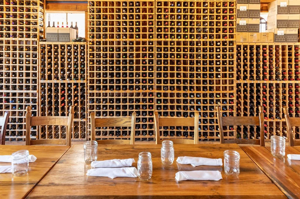A restaurant interior with a large wooden wine rack filled with bottles behind a wooden dining table set with napkins and mason jars for glasses.