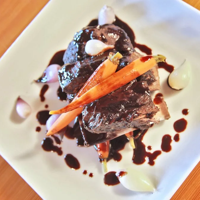 A plated dessert featuring slices of cooked meat topped with a glossy chocolate sauce, garnished with baby carrots and dollops of cream or mousse on a white square plate.