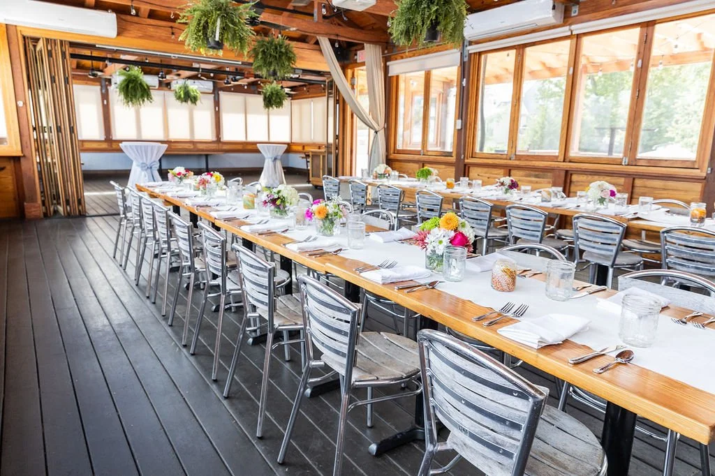Festive dining table set with flowers, glasses, and cutlery in a decorated wooden banquet hall with hanging greenery.